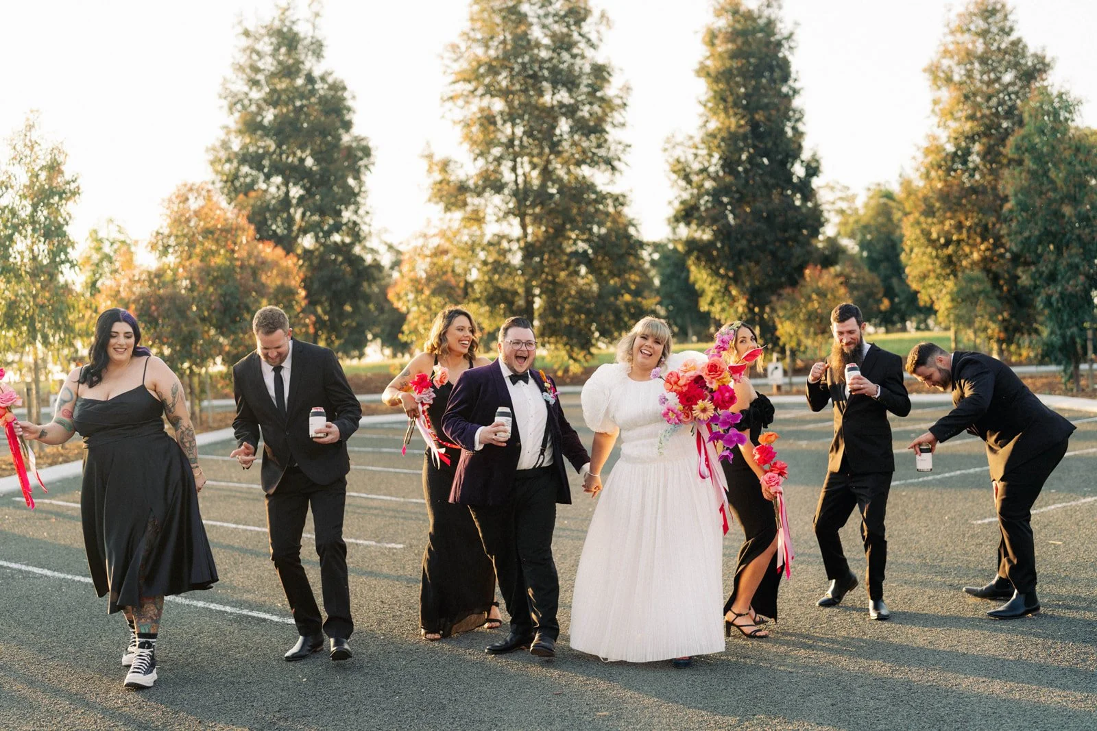 Group of people dressed in formal and casual attire, walking and laughing outdoors in a parking lot with trees and sunset in the background, holding drinks and bouquets.