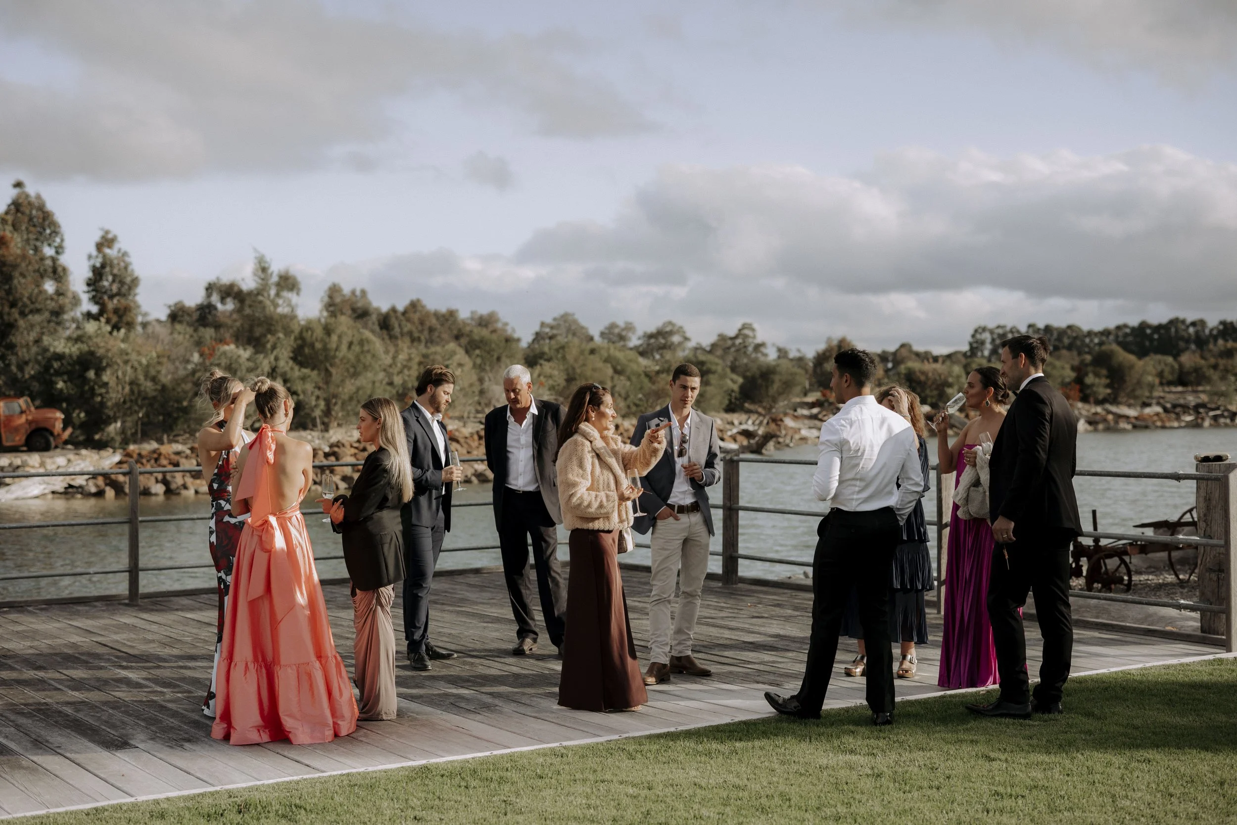 Group of men and women in formal and semi-formal attire socializing on a wooden dock by the water, some holding glasses, with trees and a cloudy sky in the background.
