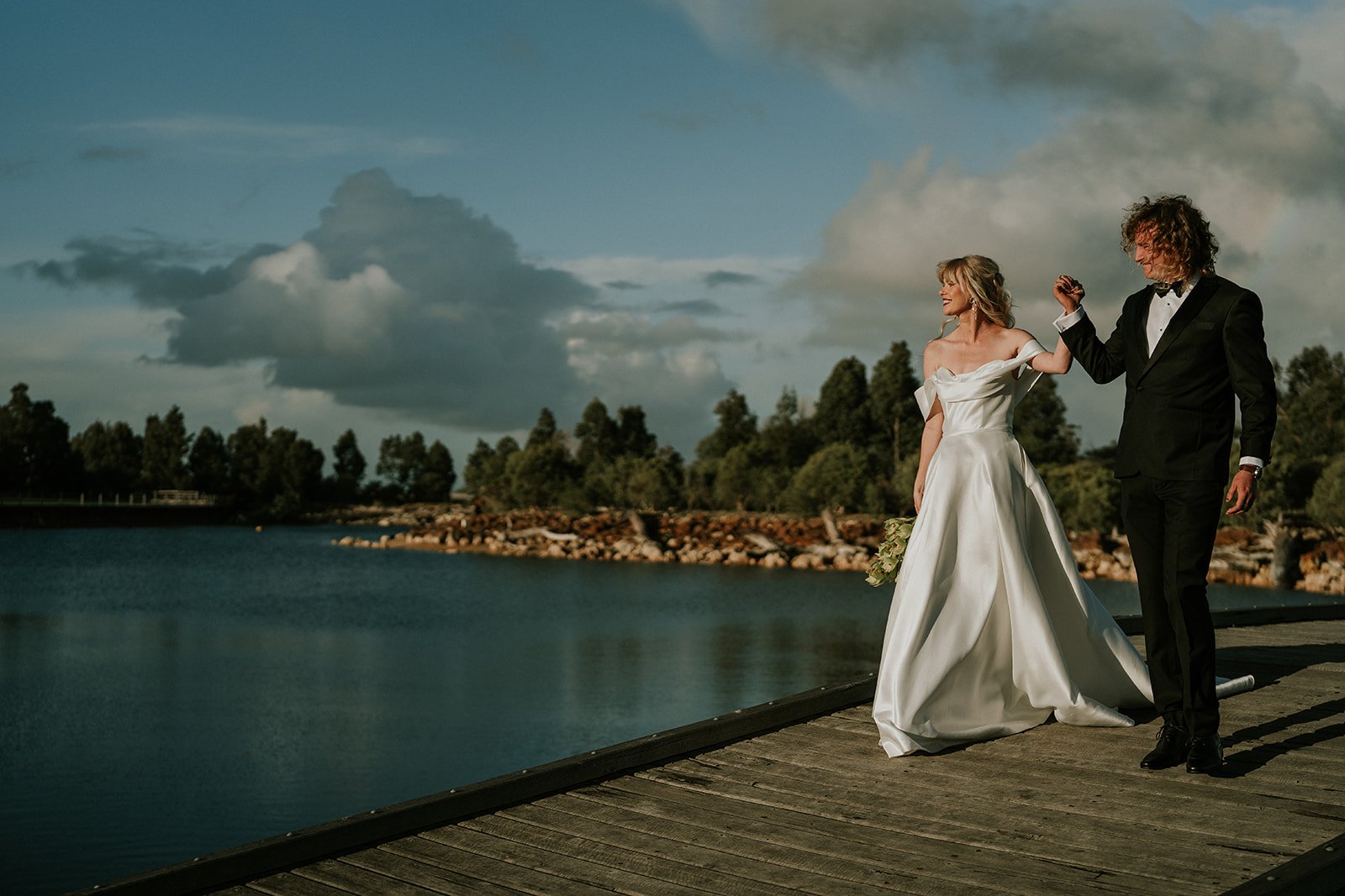 A bride and groom dancing on a wooden dock by a body of water, with trees and a cloudy sky in the background.