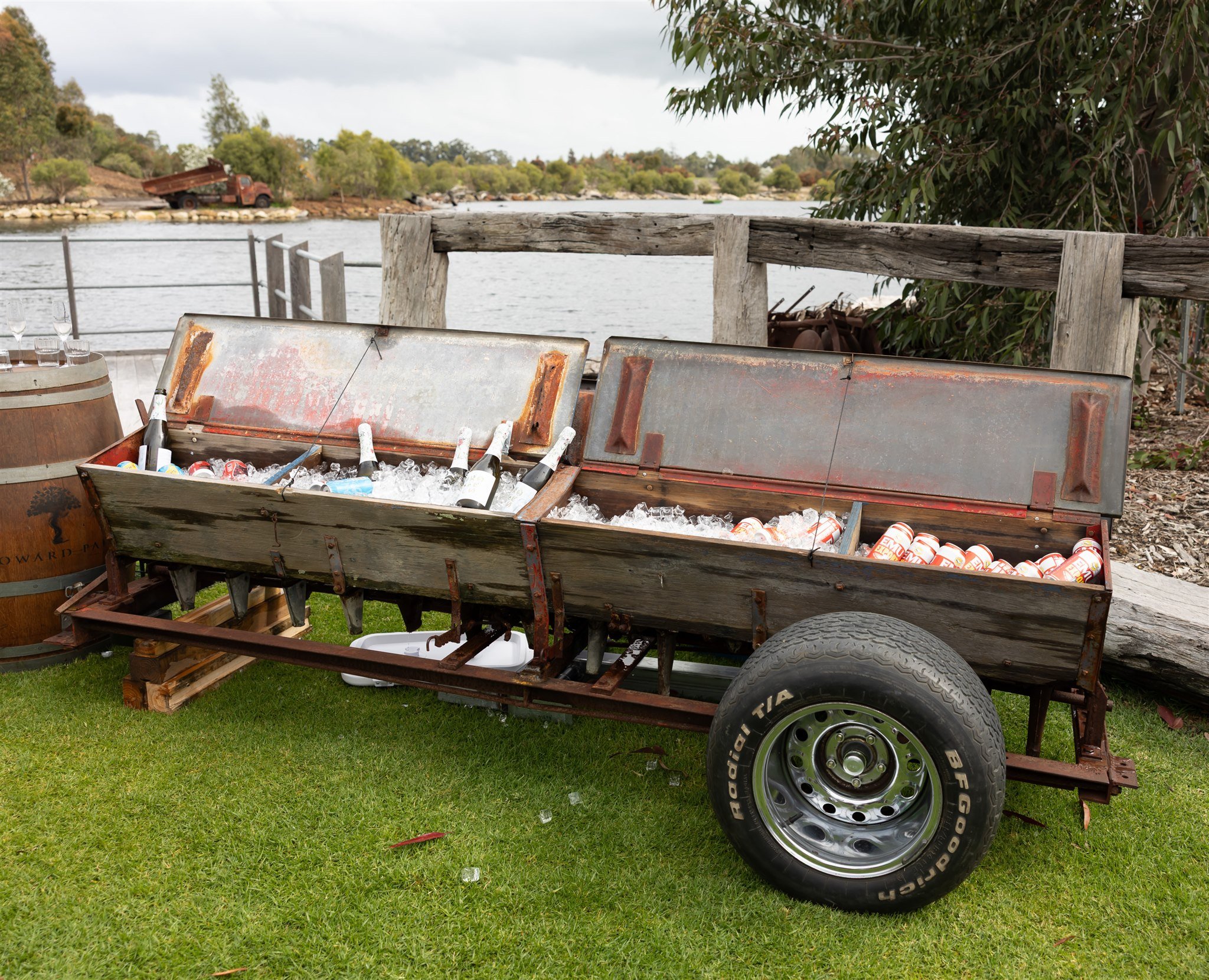 Old wooden picnic cart filled with ice and bottles of beer and soda, set outdoors near a river, with trees and a rustic fence in the background.