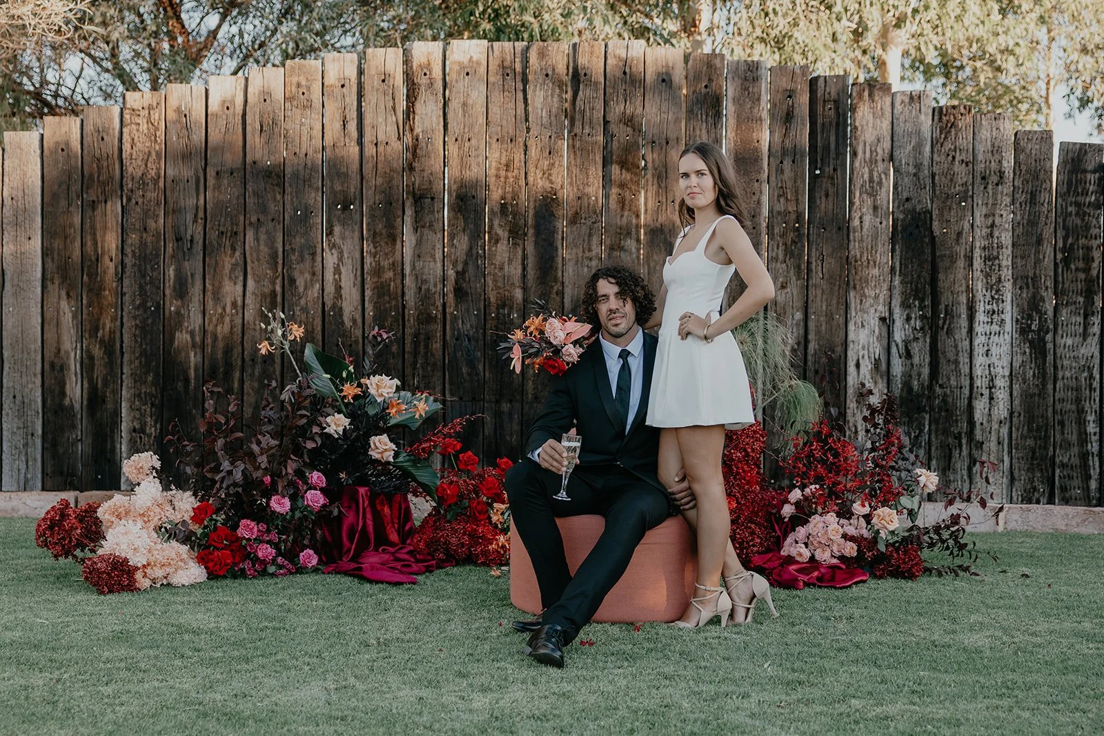 A man in a suit sitting on a pink ottoman, holding a glass of champagne, with a woman in a white dress standing beside him with one foot on his knee, in front of a wooden fence and floral decorations.