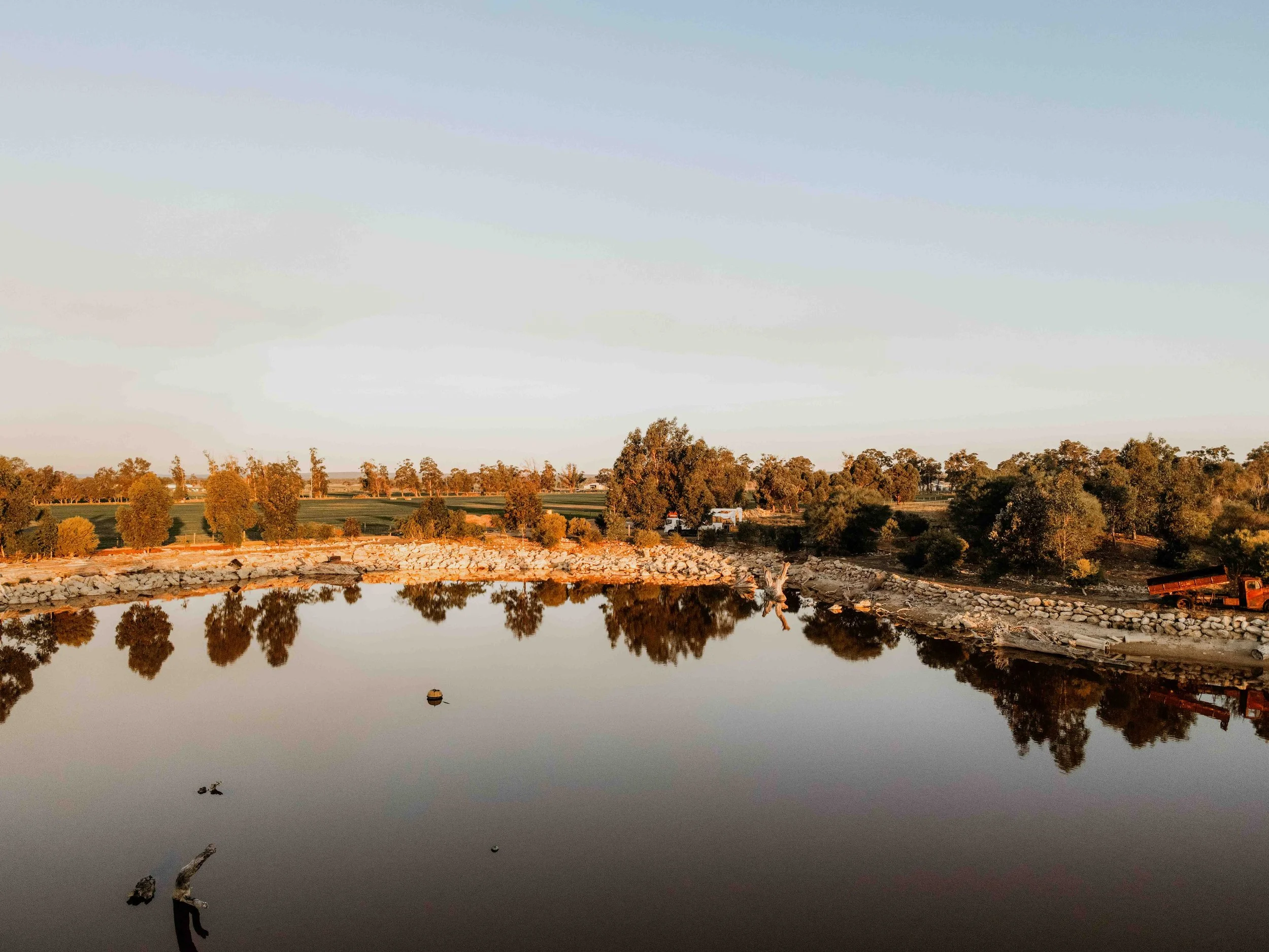 A calm body of water reflecting trees and a clear sky during sunset, with some rocks and a boat on the shoreline.