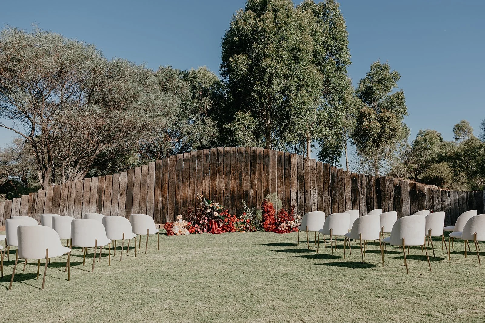 Outdoor wedding ceremony setup with white chairs arranged in rows on a grassy area, floral arrangements at the front, wooden privacy fence, tall trees in the background, and a clear blue sky.