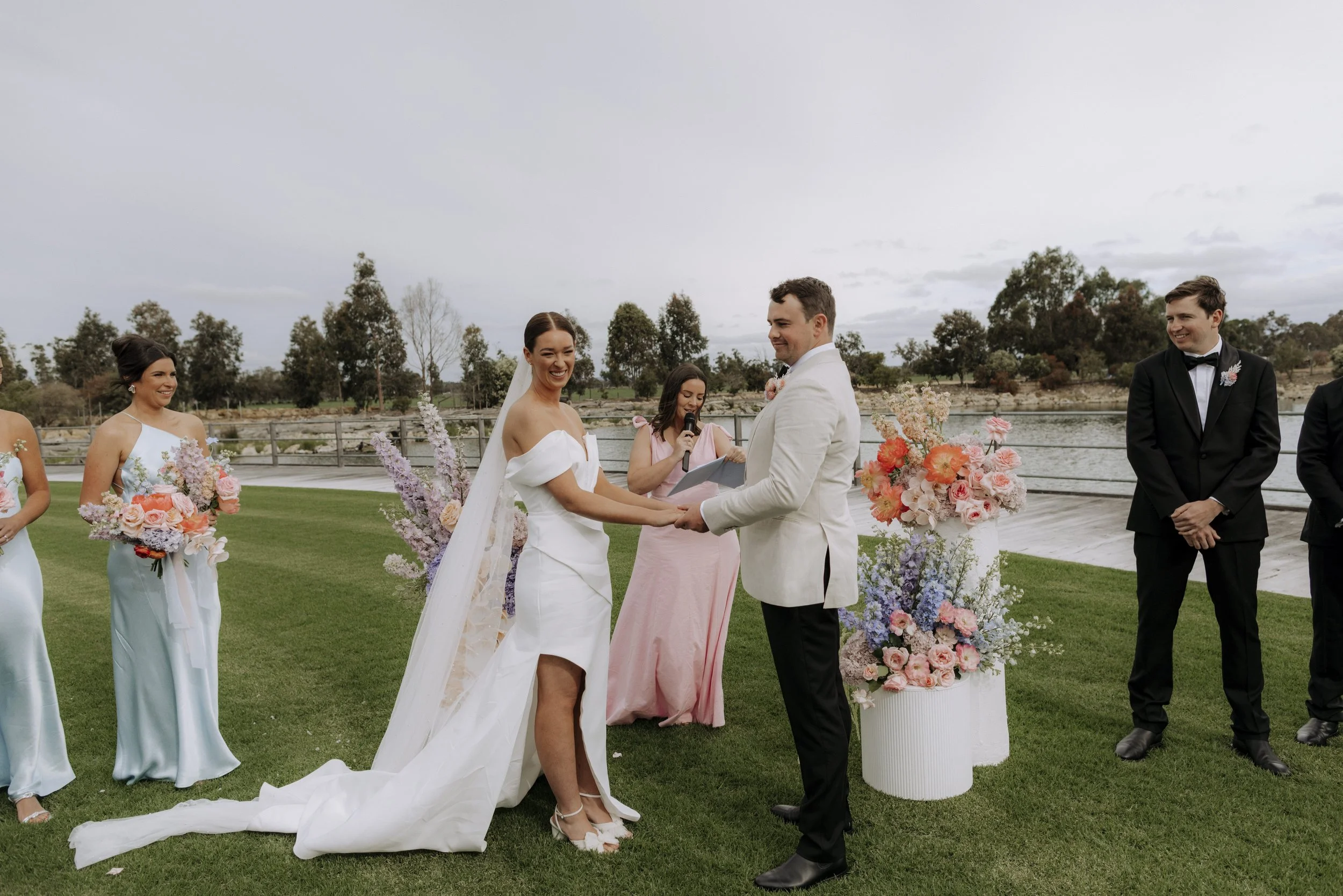 A wedding ceremony outdoors by a lake with seven people, including a bride in a white off-shoulder wedding dress, a groom in a white tuxedo jacket, four bridesmaids in matching pastel blue dresses holding bouquets, and a groomsman in a black tuxedo, with floral arrangements and a scenic backdrop of water and trees.