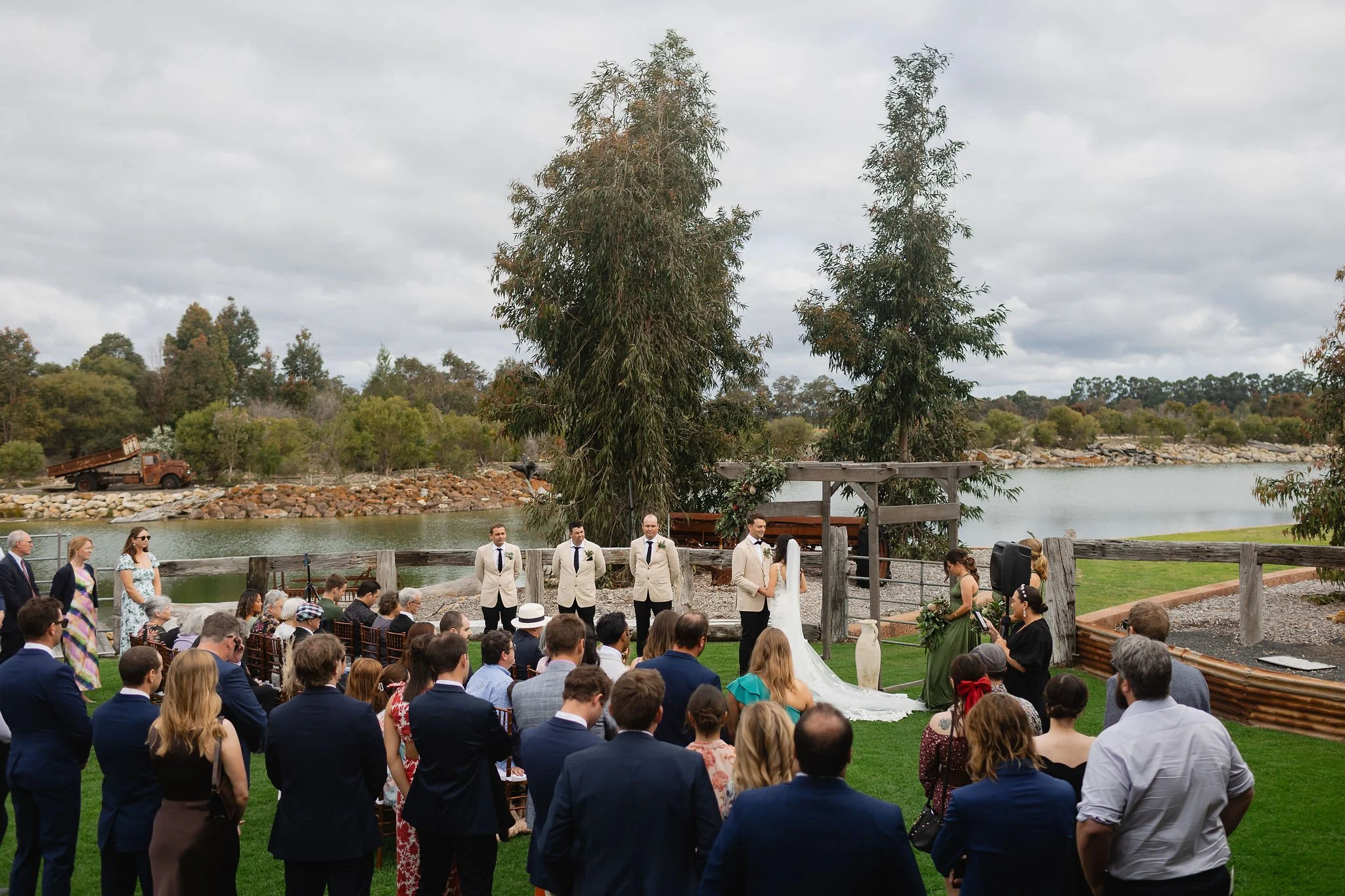 Outdoor wedding ceremony by a lake with guests standing on the grass, a bride and groom at the altar, and officiants alongside. Overcast sky, trees, boat, and lake in the background.