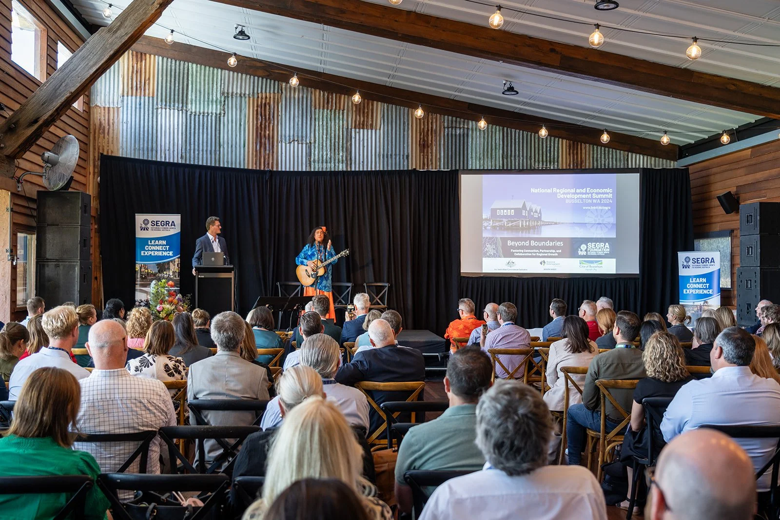 A speaker is playing guitar on a stage at a conference, with a large audience seated and listening. The conference banner reads 'National Regional and Economic Development Summit BUSSELOW WA 2024'.