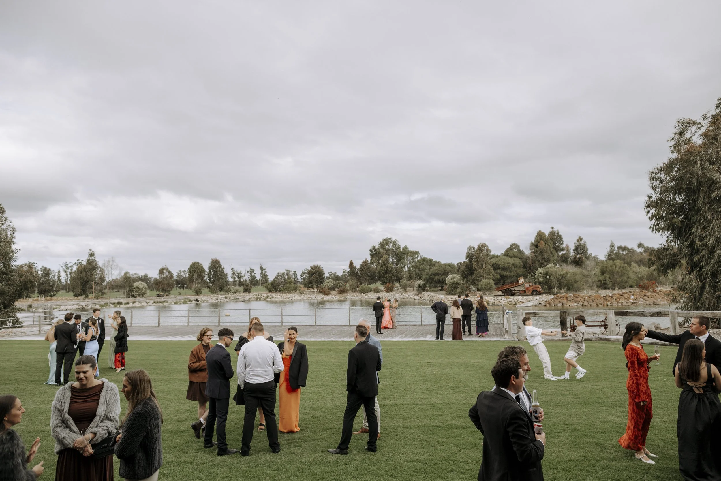 People dressed in formal attire socializing on a grassy area near a river, with some engaging in conversation and others taking photos under a cloudy sky.