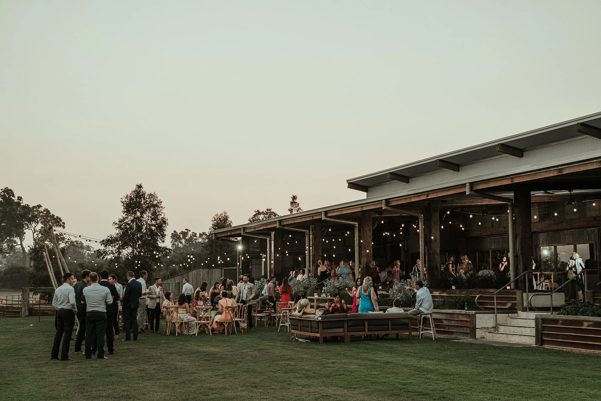 A large outdoor gathering with people socializing and sitting at tables on grass in front of a wooden building decorated with string lights, during dusk