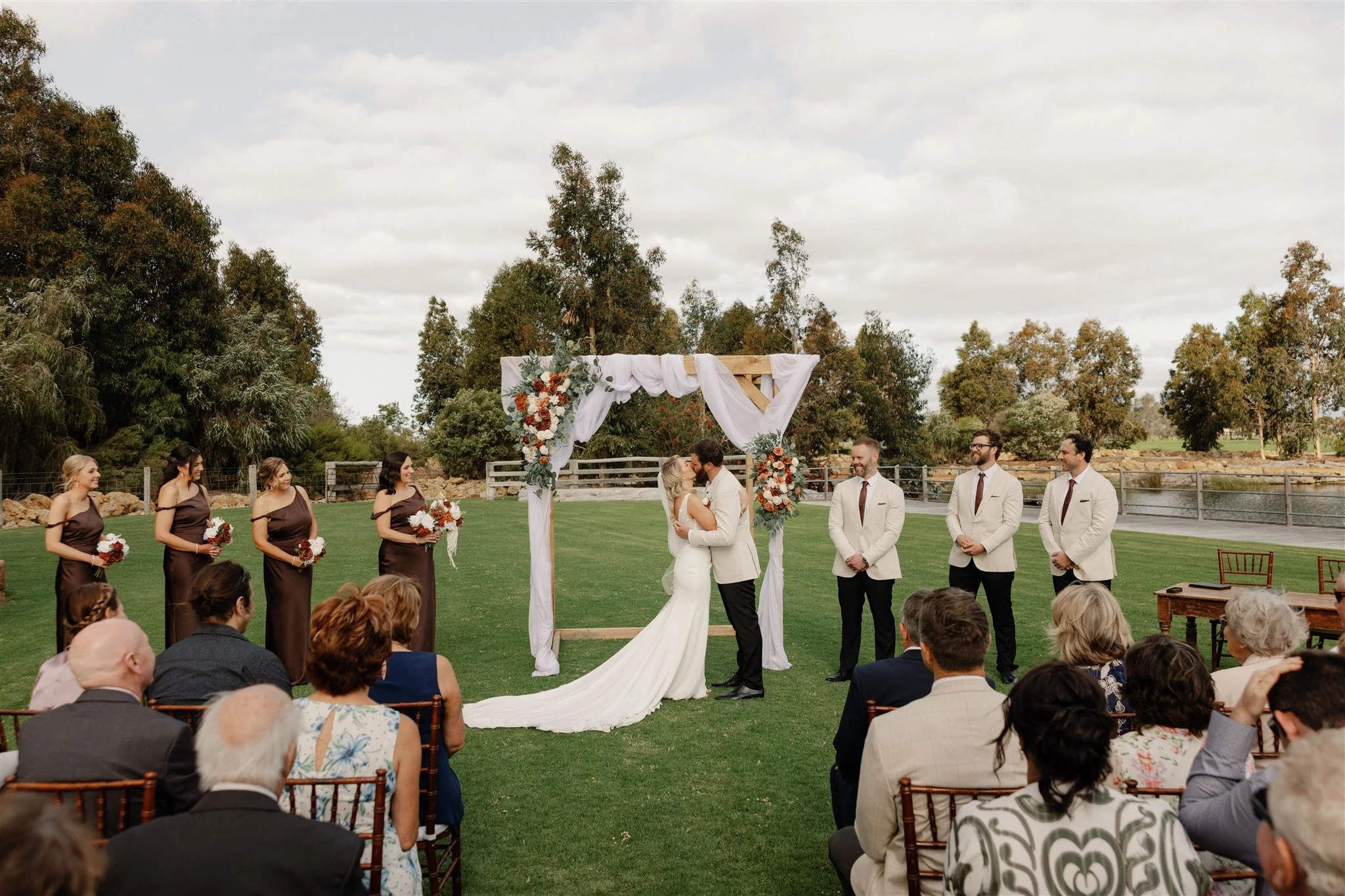 Bride and groom exchanging vows at outdoor wedding ceremony under an arch decorated with white fabric and floral arrangements, with bridesmaids and groomsmen standing on either side, and guests seated watching.