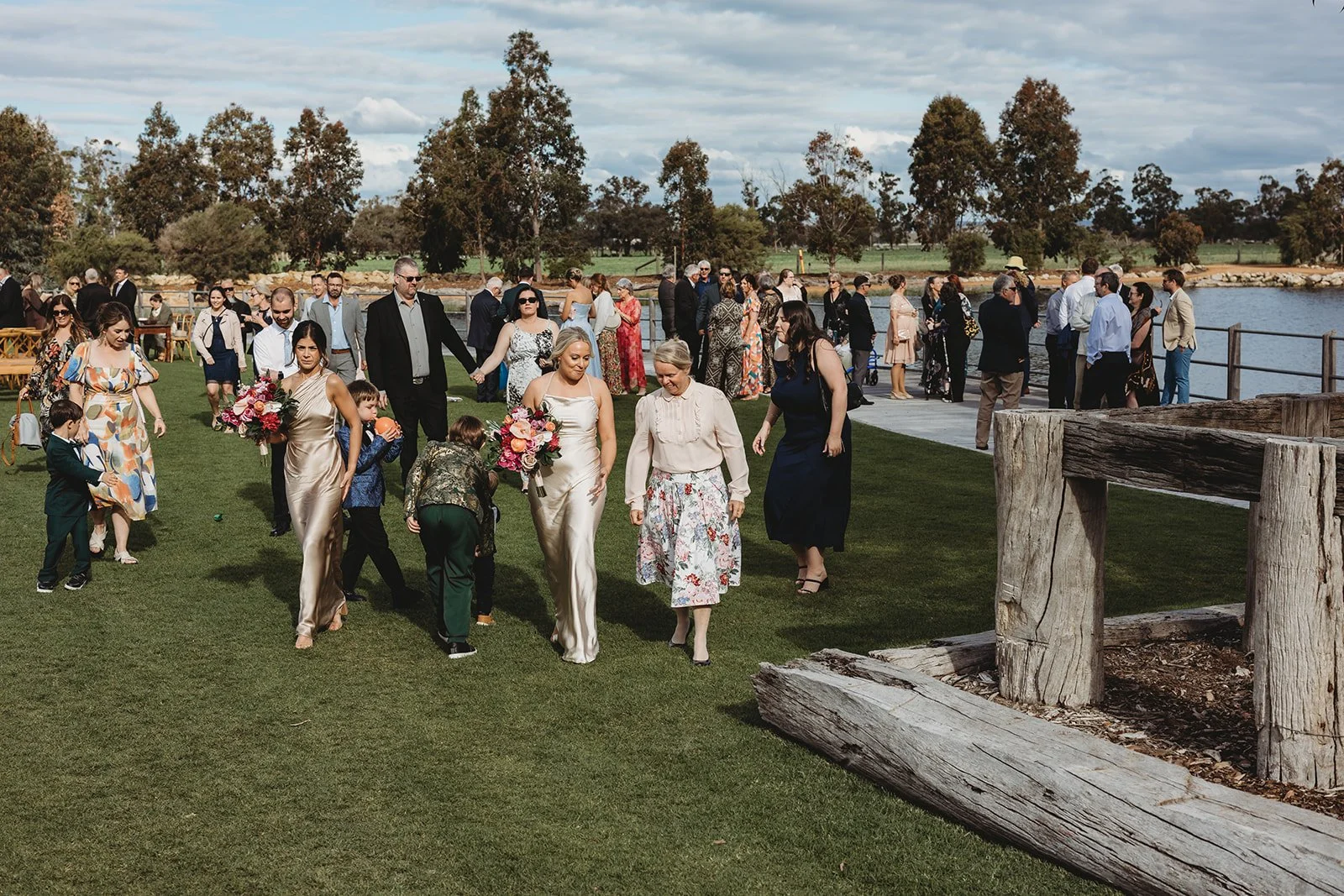 People gathered outdoors by a lake for a wedding celebration, including women in formal dresses, children, and men in suits, with trees and a cloudy sky in the background.