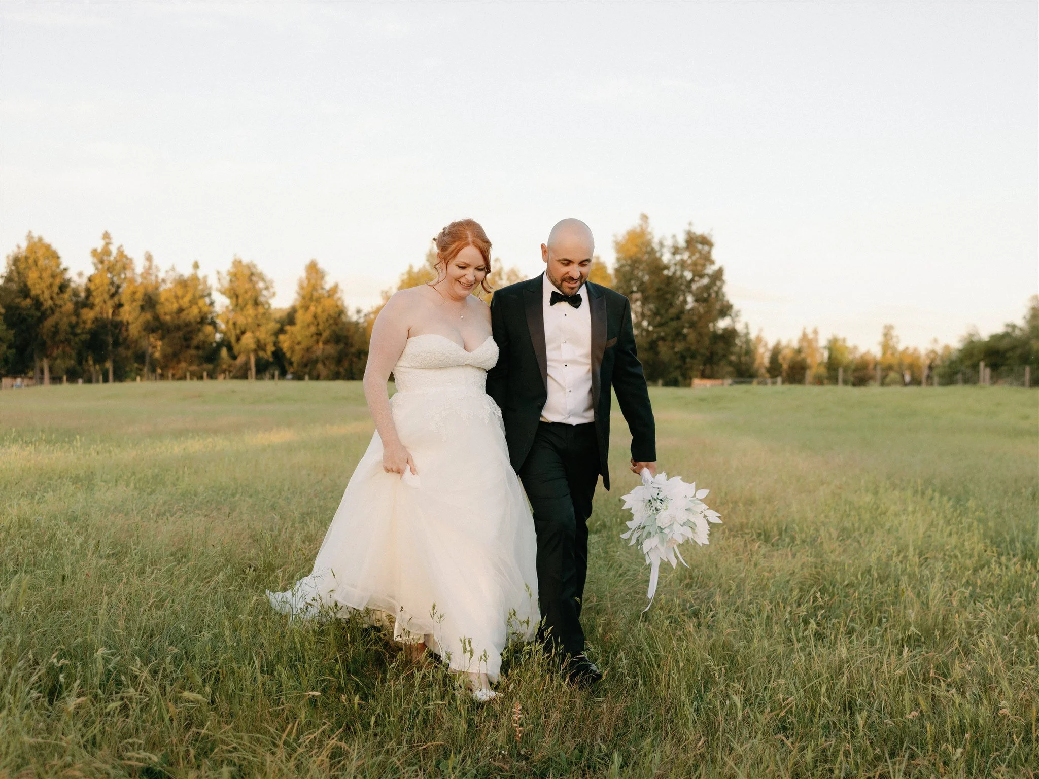 A bride and groom walking hand in hand through a grassy field, with the bride holding her wedding dress with her left hand and the groom holding a white bouquet in his right hand, surrounded by trees under a clear sky.