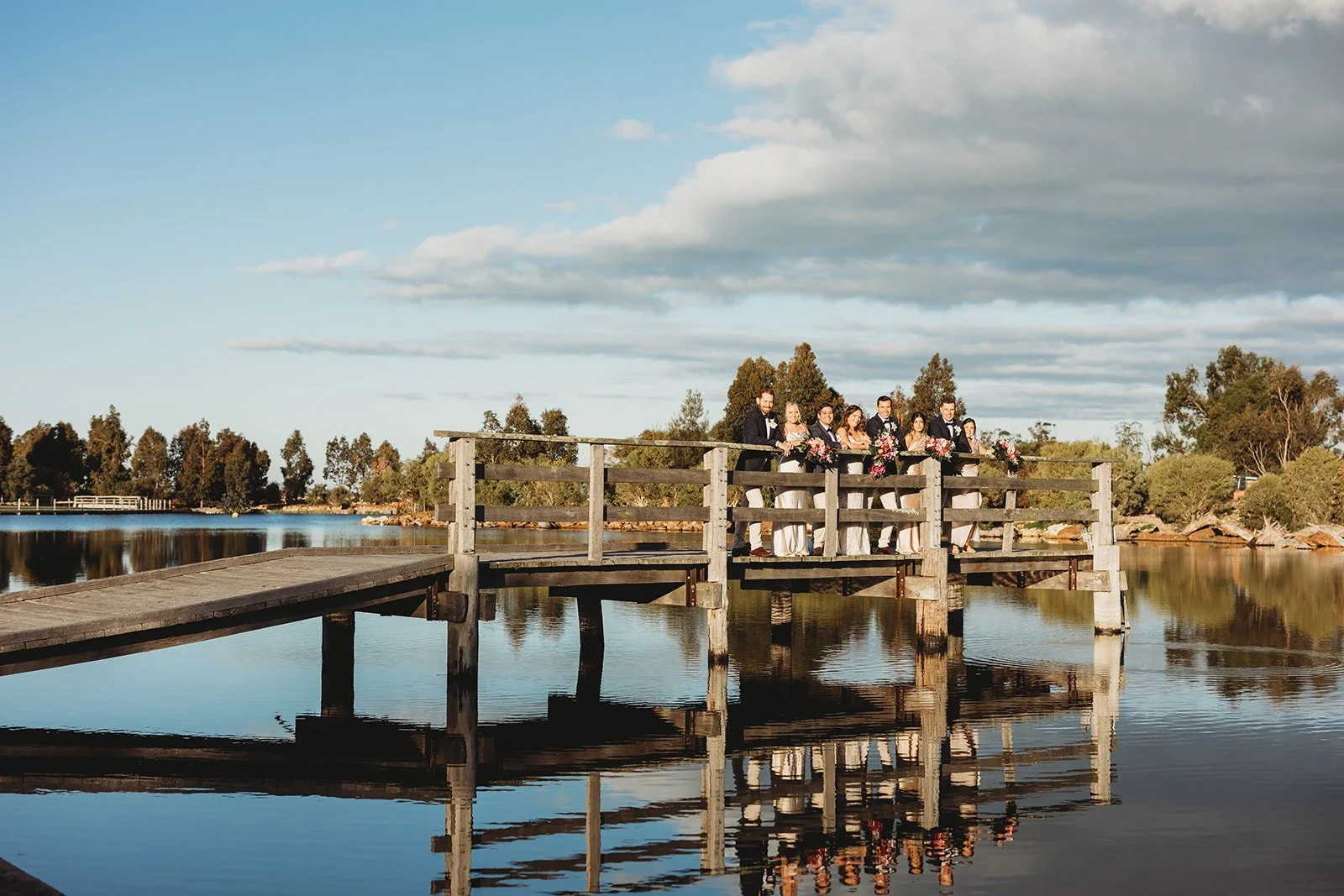 A wedding party standing on a wooden dock over a calm lake during sunset, with trees and a blue sky with clouds in the background.