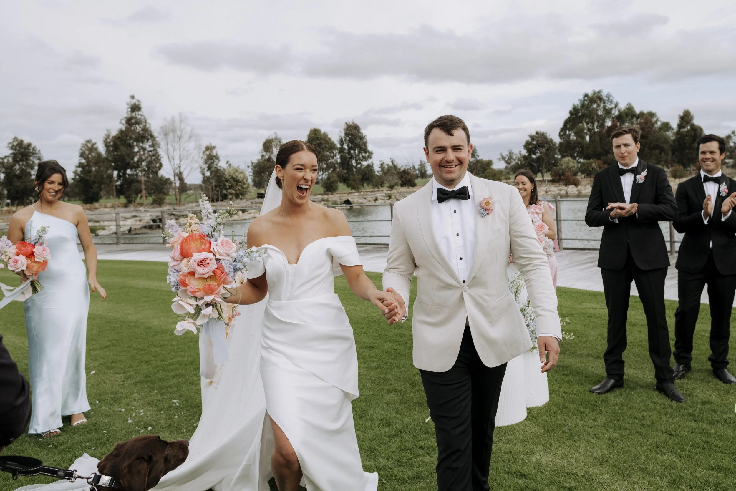 A newlywed couple walking hand in hand outdoors, smiling and celebrating, with wedding guests clapping and smiling in the background near a lake, on a cloudy day.