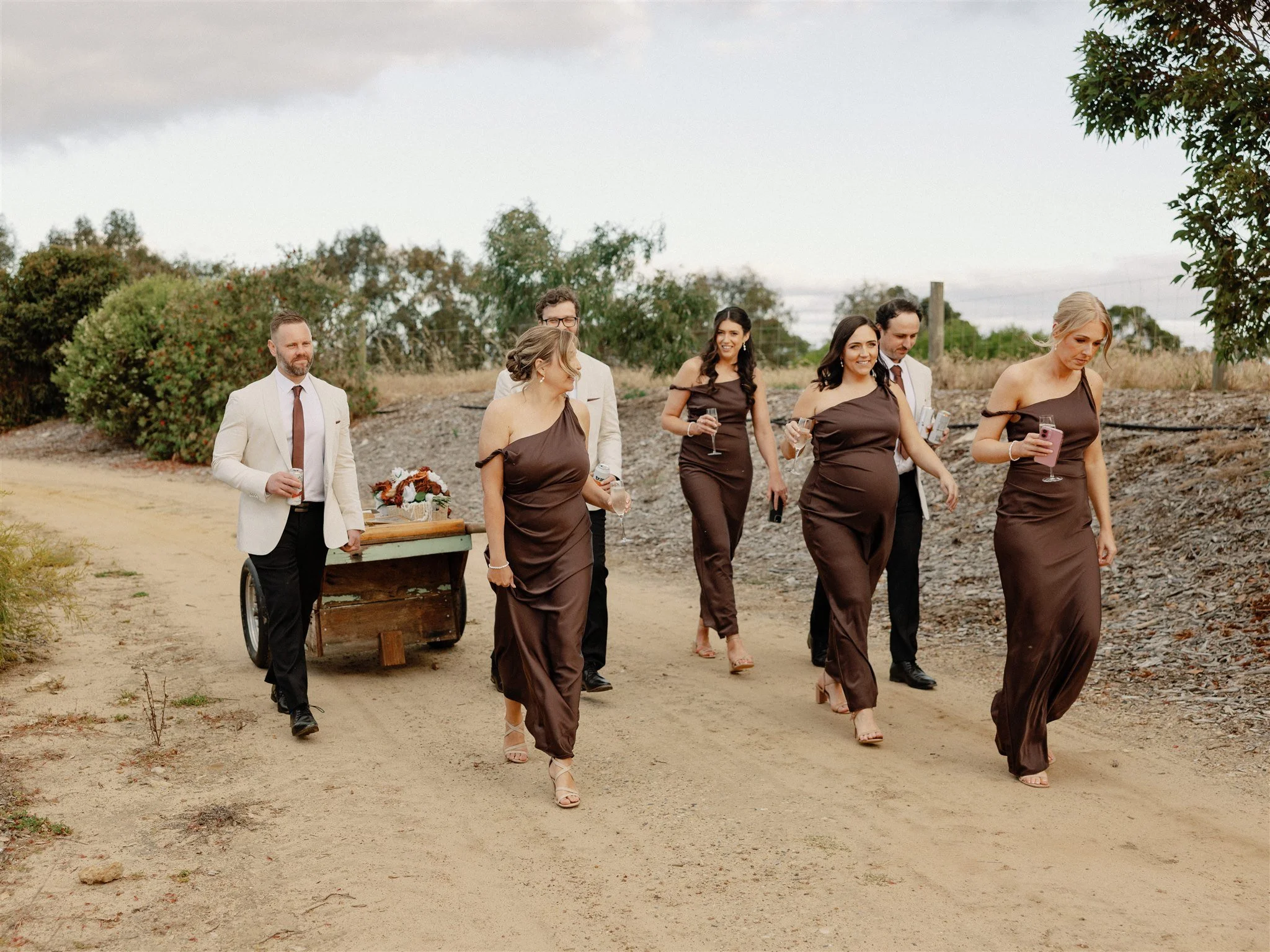 Group of seven people dressed in formal attire walking on a dirt path outdoors, some holding drinks, with trees and cloudy sky in the background.