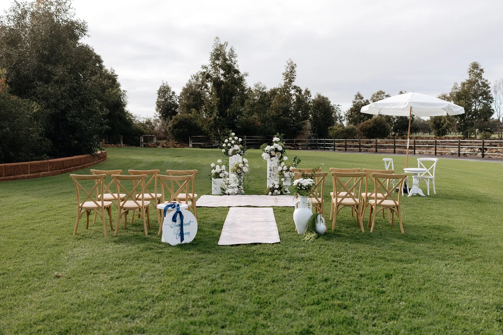 Outdoor wedding setup on a grassy field with floral arrangements, chairs, and a white umbrella.