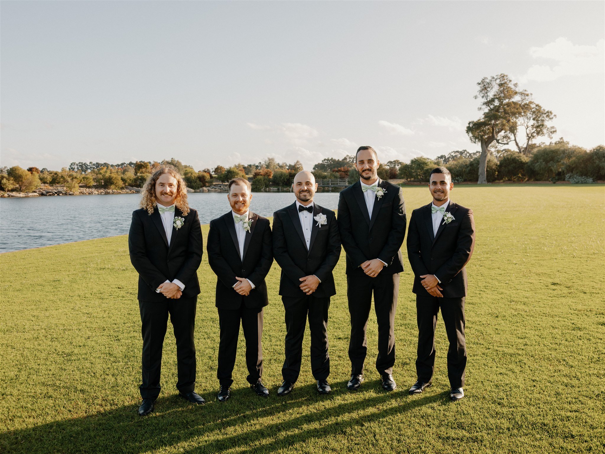 Group of five men in tuxedos standing on grass by a lake at sunset.