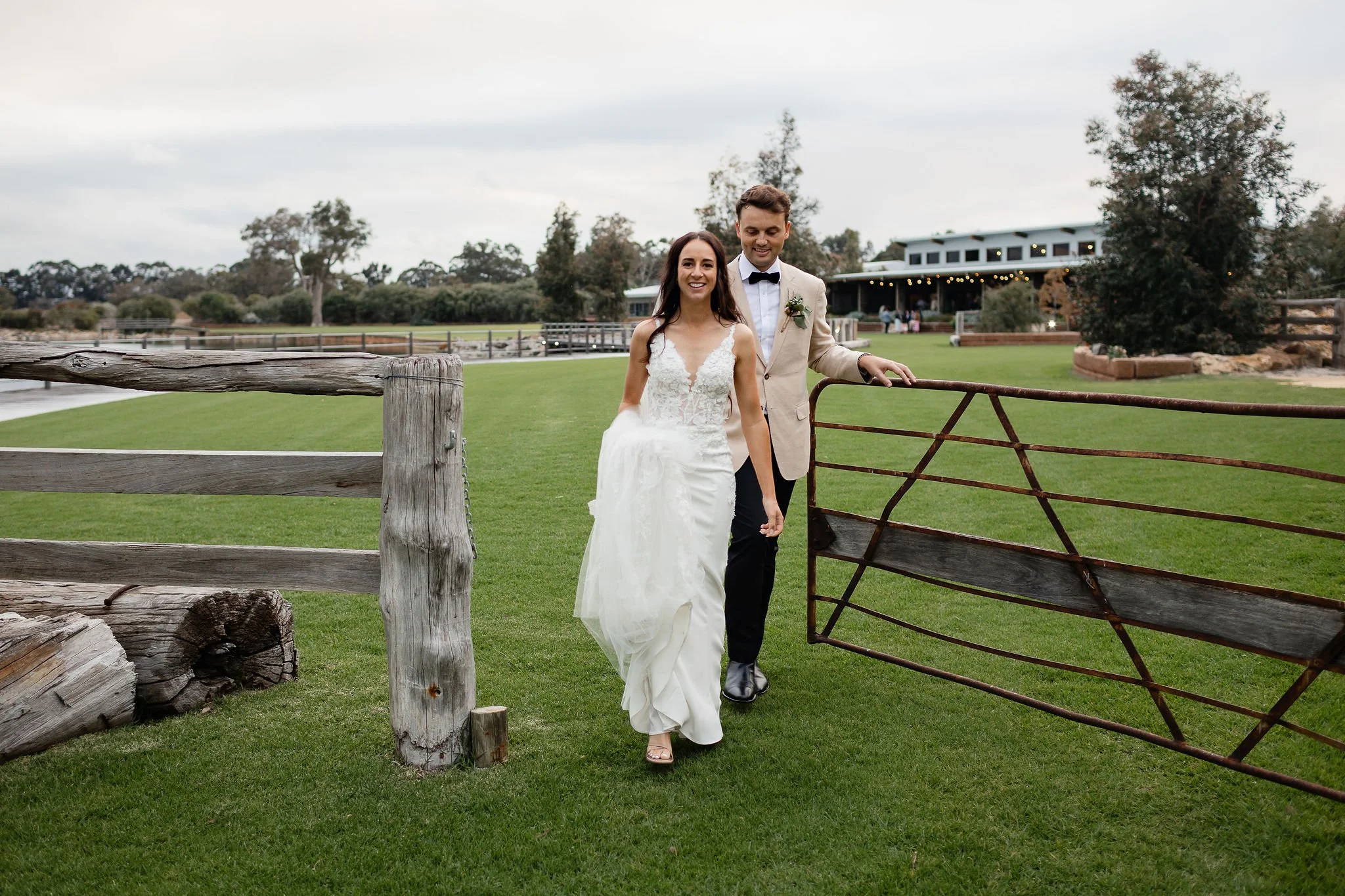 A bride and groom walking through a rustic outdoor wedding venue with green grass, trees, and a barn in the background.