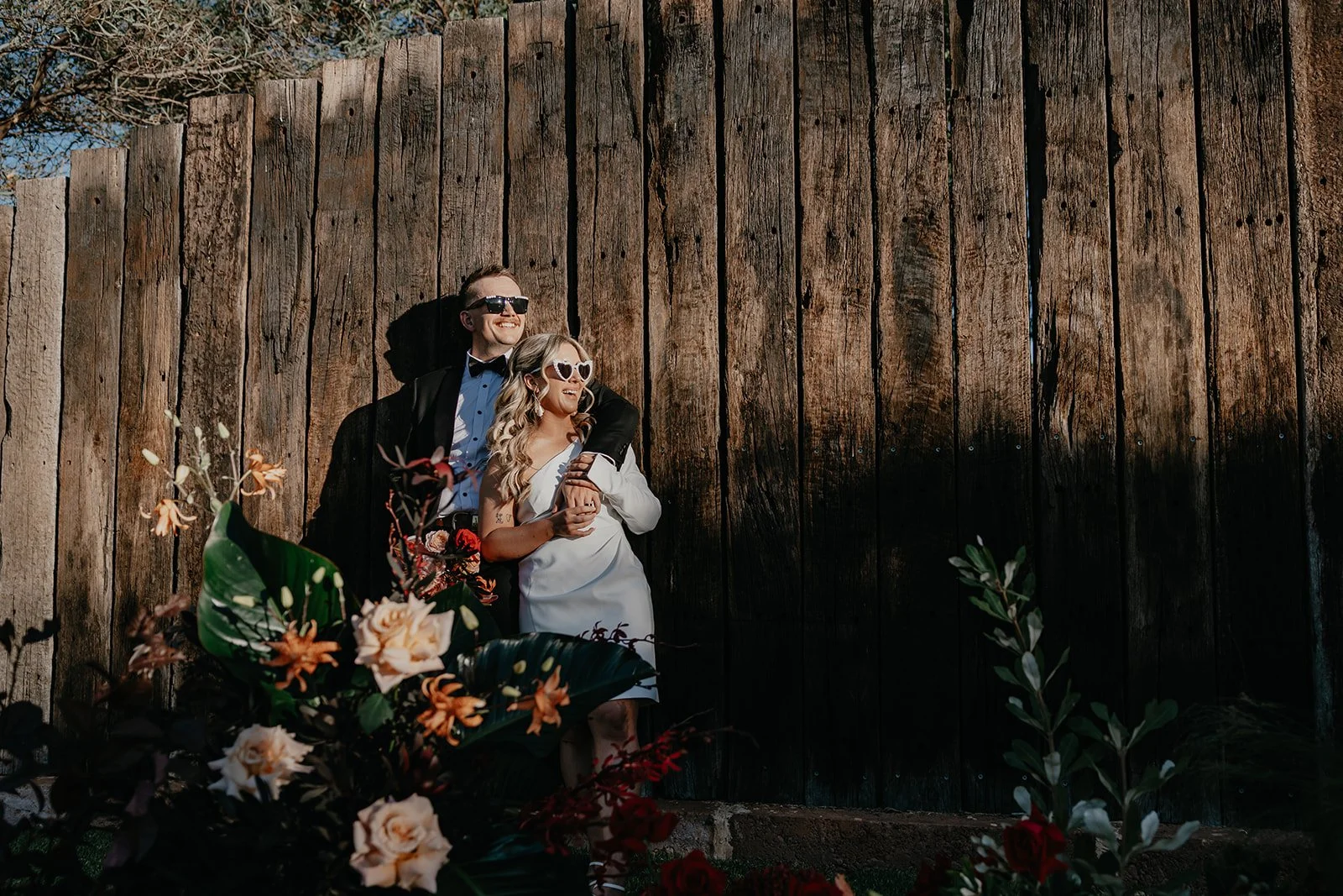 A couple dressed in formal attire leaning against a tall wooden fence with flowers in the foreground. The man is wearing a black tuxedo and sunglasses, and the woman is wearing a white dress and sunglasses, smiling and looking to the side.