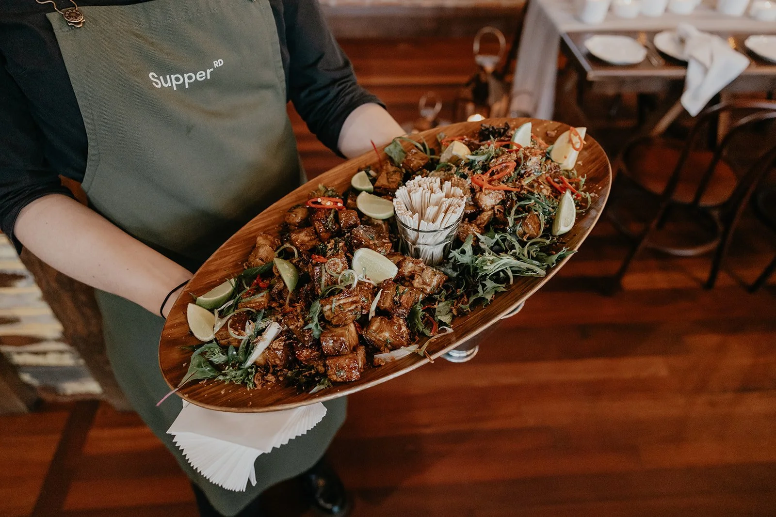 Waitstaff holding a large wooden platter filled with assorted grilled meats, lime wedges, and garnishes at a restaurant.