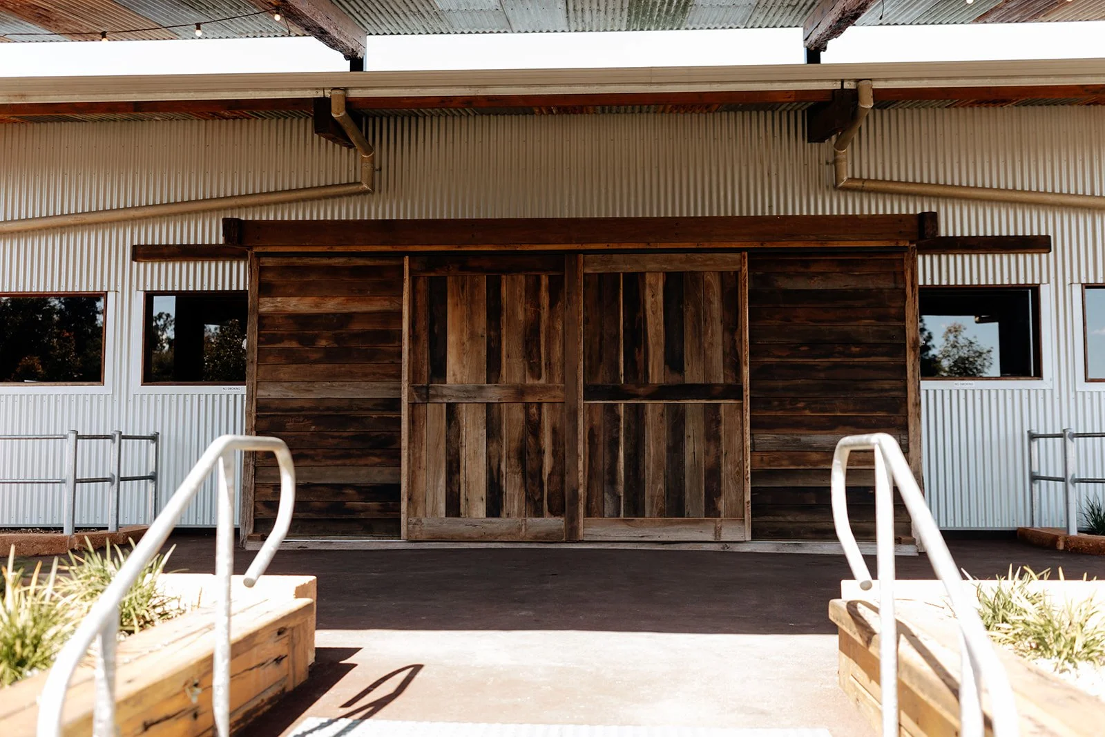 Wooden barn door on a building with metal siding, with stairs and railings leading up to it, and planters with plants in the foreground.