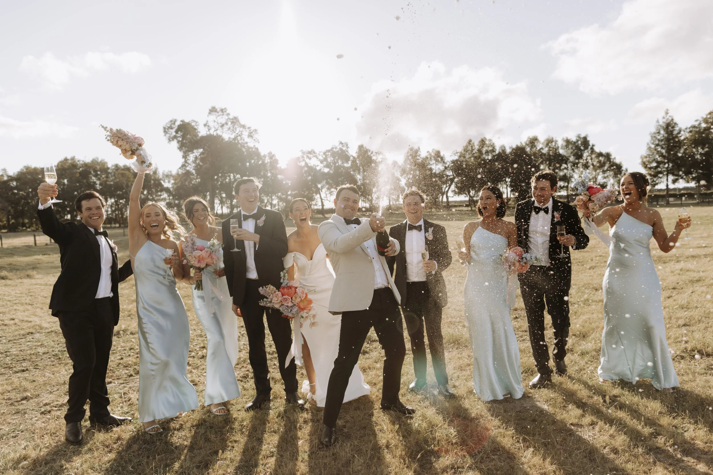 Wedding celebration outdoors with smiling bride, groom, bridesmaids, groomsmen, and guests, with some holding champagne glasses and bouquets, sunny sky and trees in background.