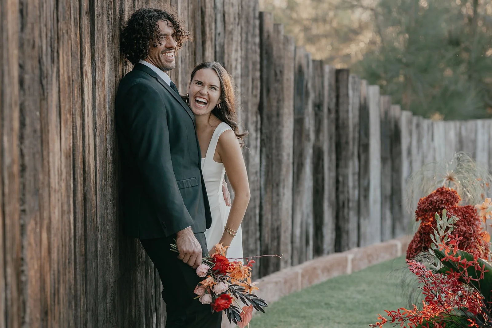 A couple dressed in wedding attire leaning against a wooden fence outdoors, with the man holding a bouquet and both smiling and laughing.