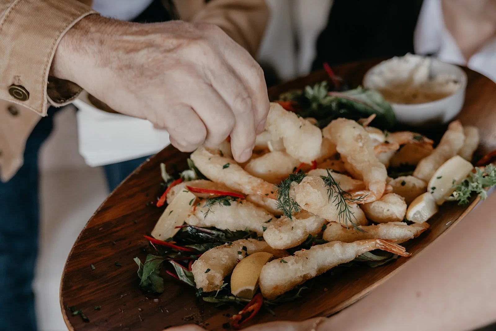Person's hand placing fried fish fillets on a wooden platter garnished with herbs, lemon wedges, and vegetables, with dipping sauce in a small bowl in the background.