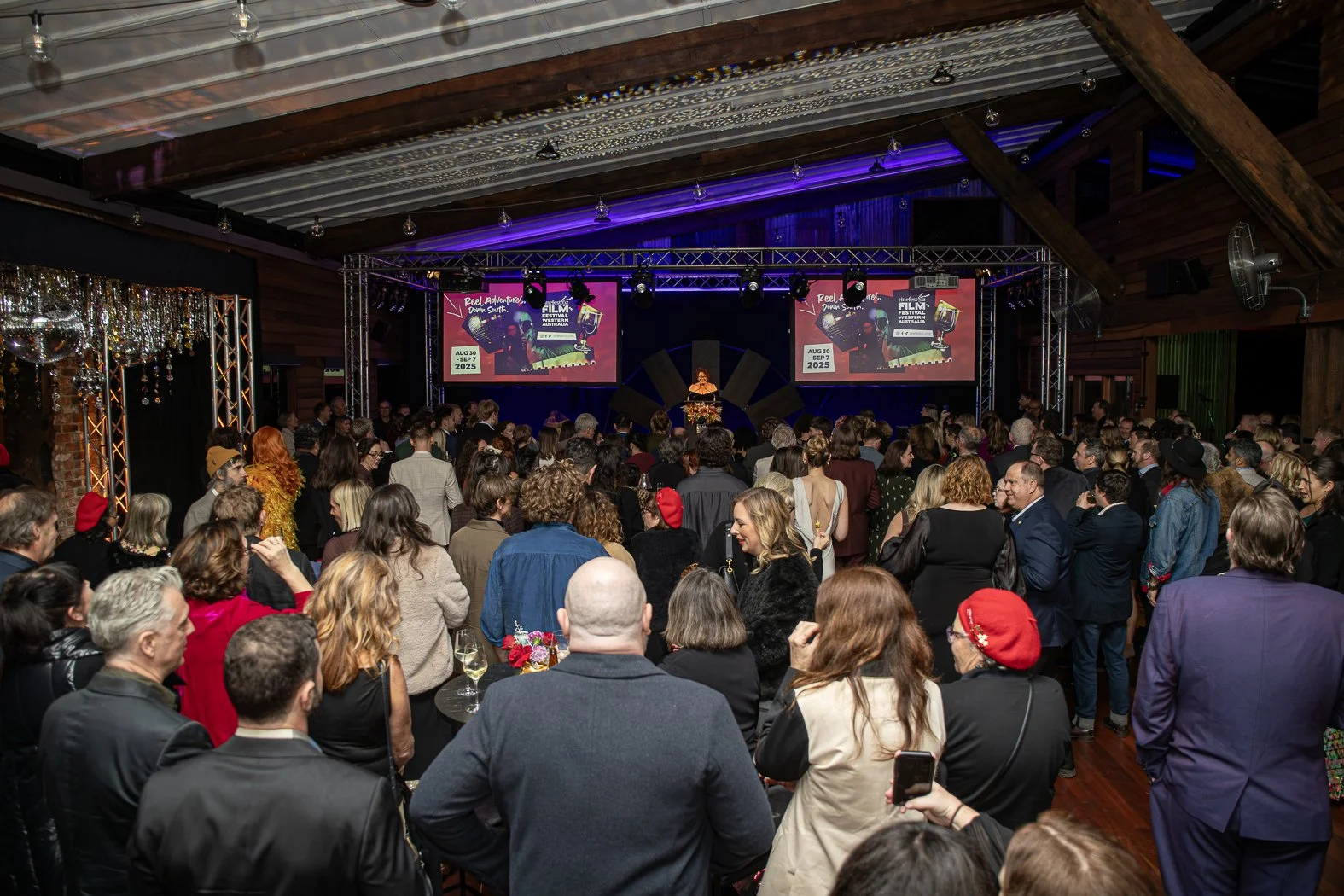 Crowd attending a film festival event in a large indoor venue with a stage and multiple screens displaying promotional material.