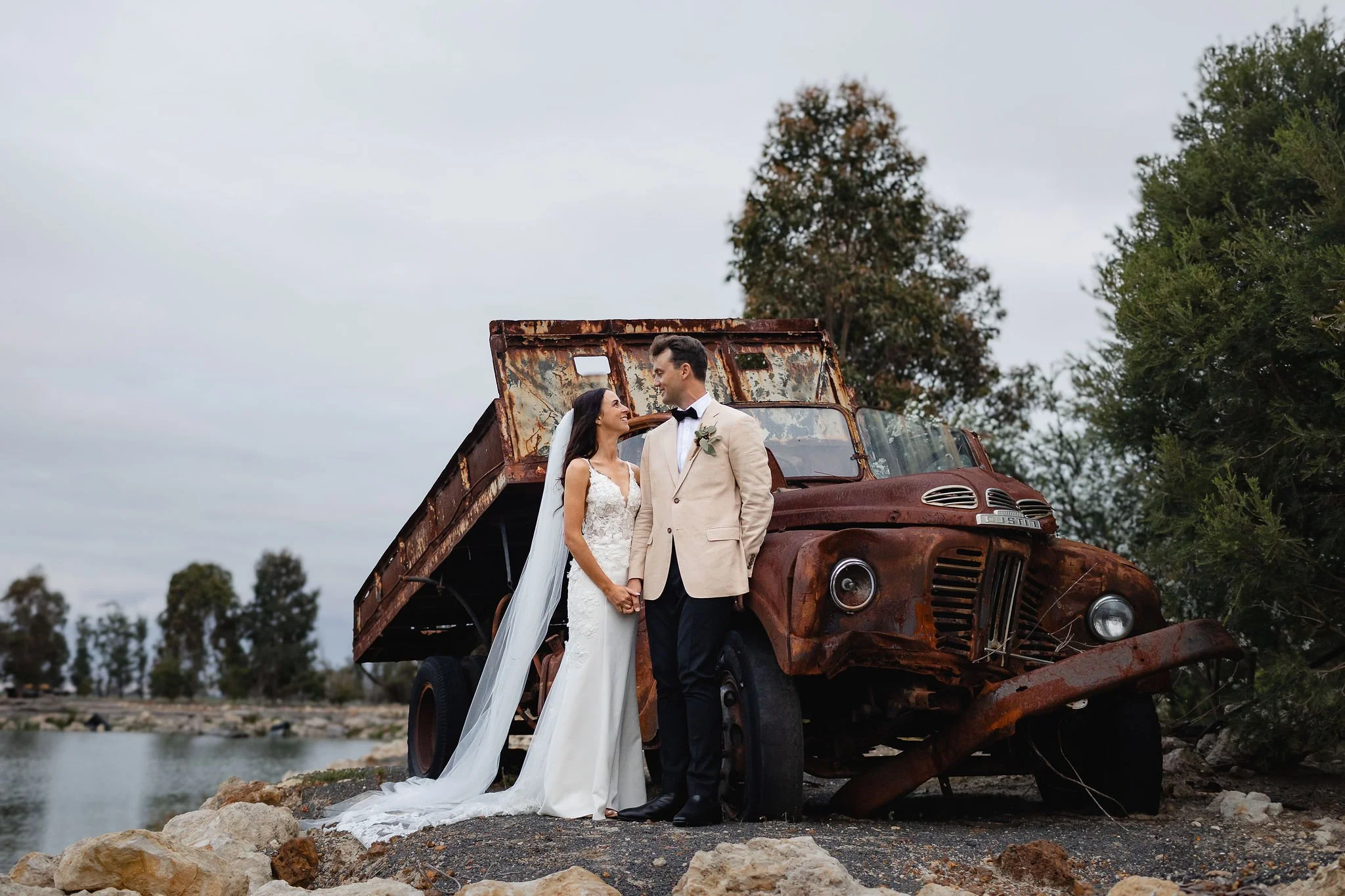 A bride and groom in wedding attire standing hand in hand, smiling at each other, next to a rusty, abandoned truck near a body of water, with trees and cloudy sky in the background.