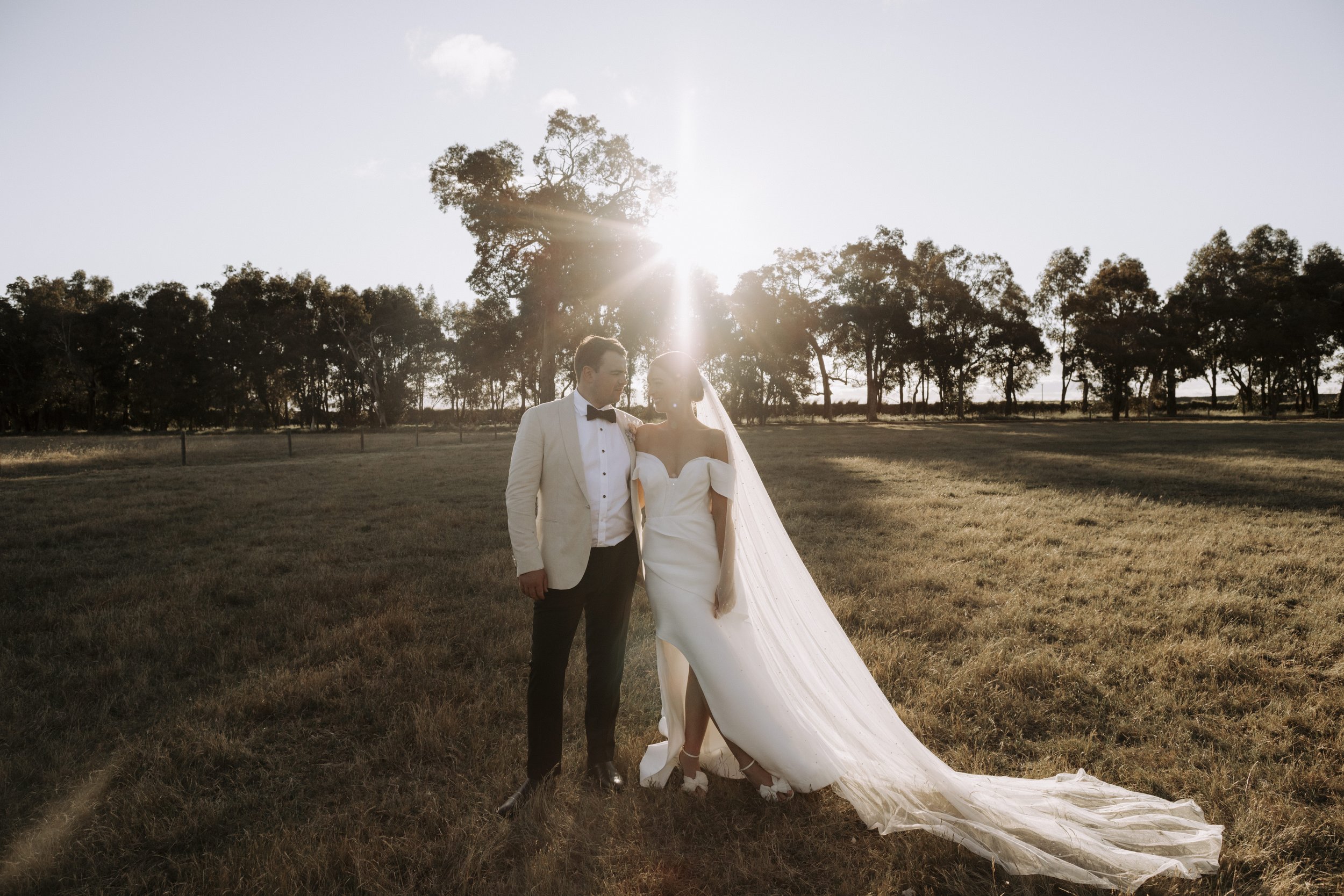A bride and groom in wedding attire standing in a field with trees in the background, backlit by the setting sun.
