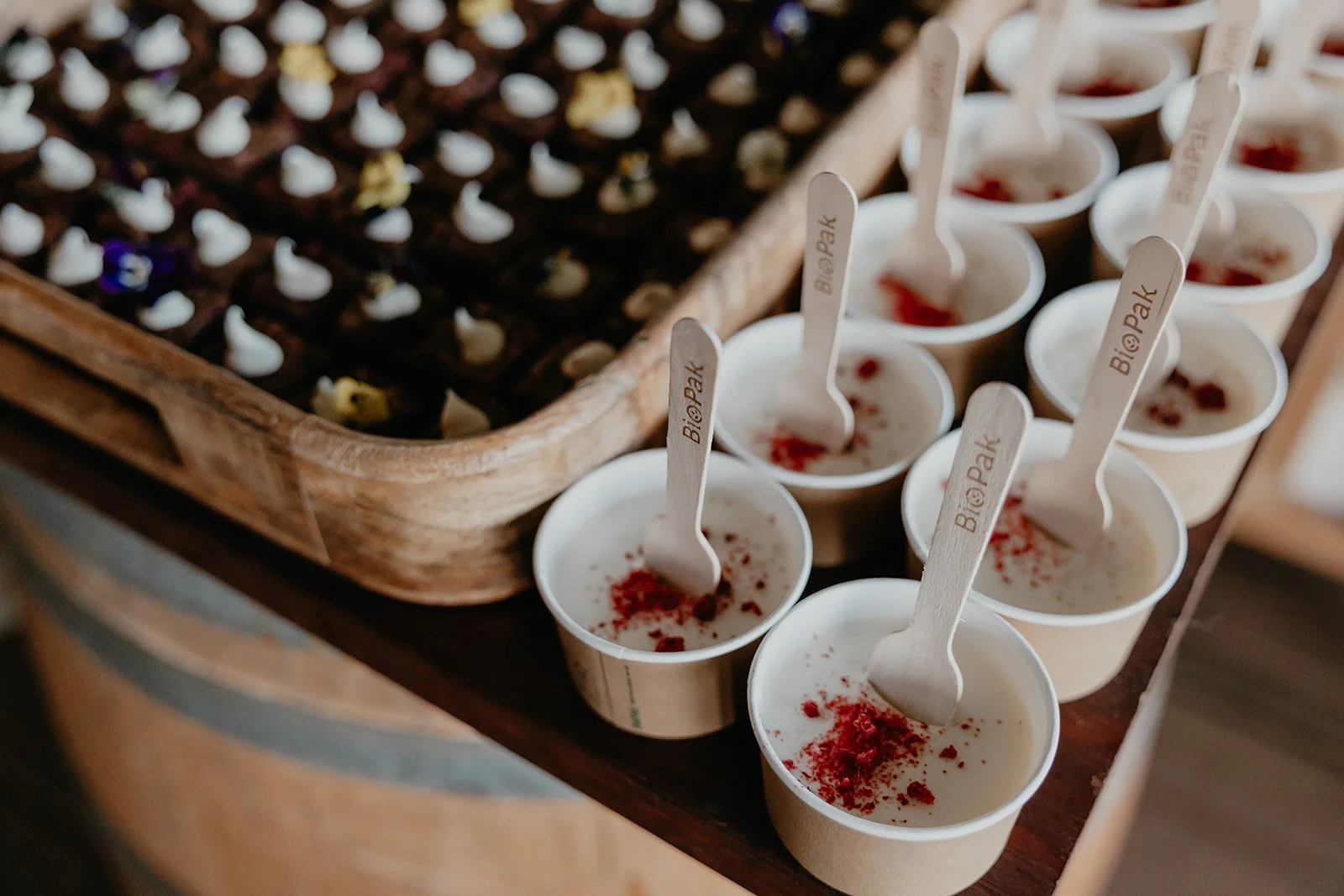 Multiple small cups of creamy dessert with red topping and wooden sticks with 'Big @ Park' printed on them, arranged on a wooden surface, with a wooden tray of chocolates in the background.