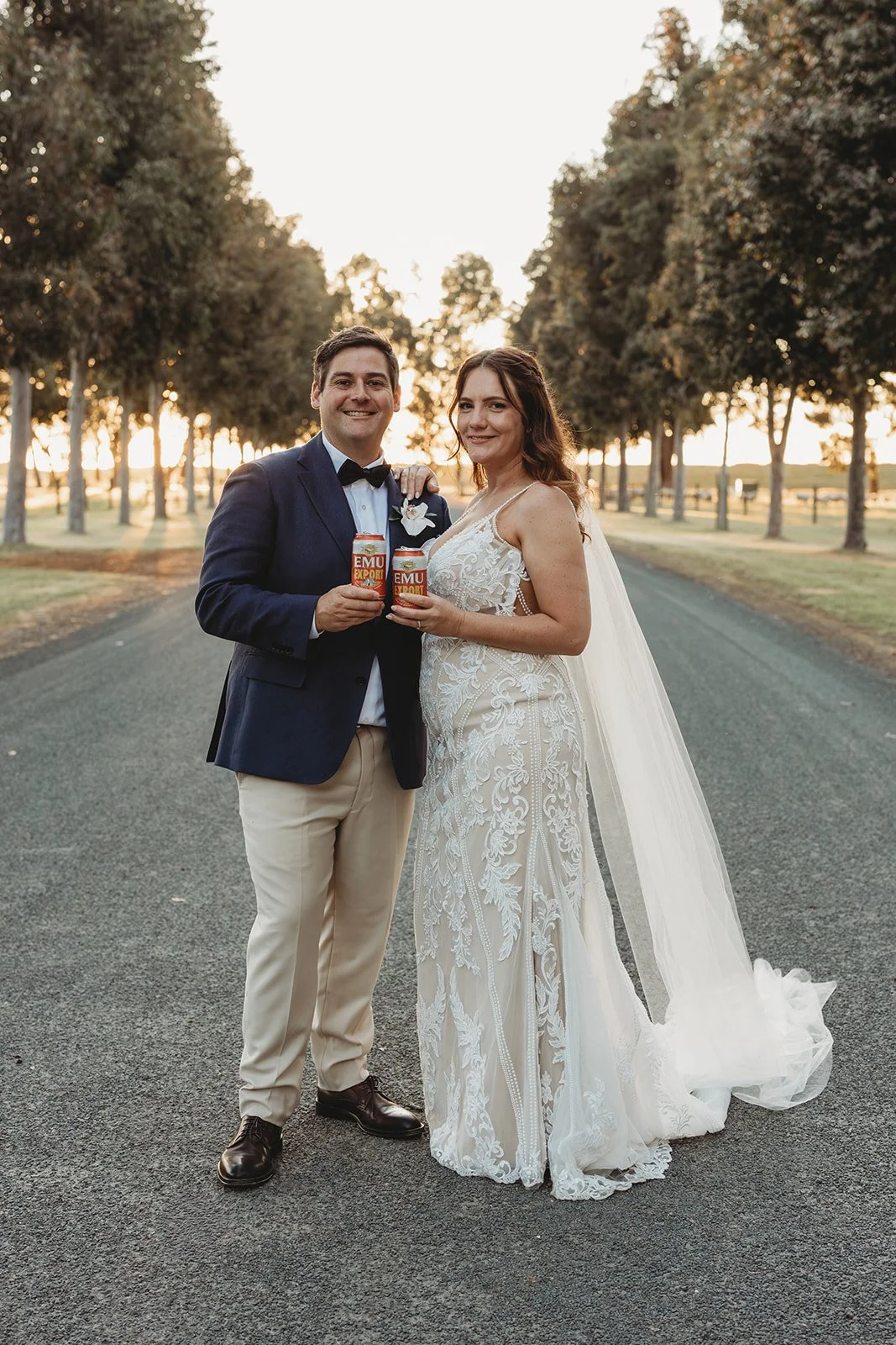 A newlywed couple in wedding attire standing on an empty road lined with trees during sunset, holding cans of EMU export beer and smiling at the camera.