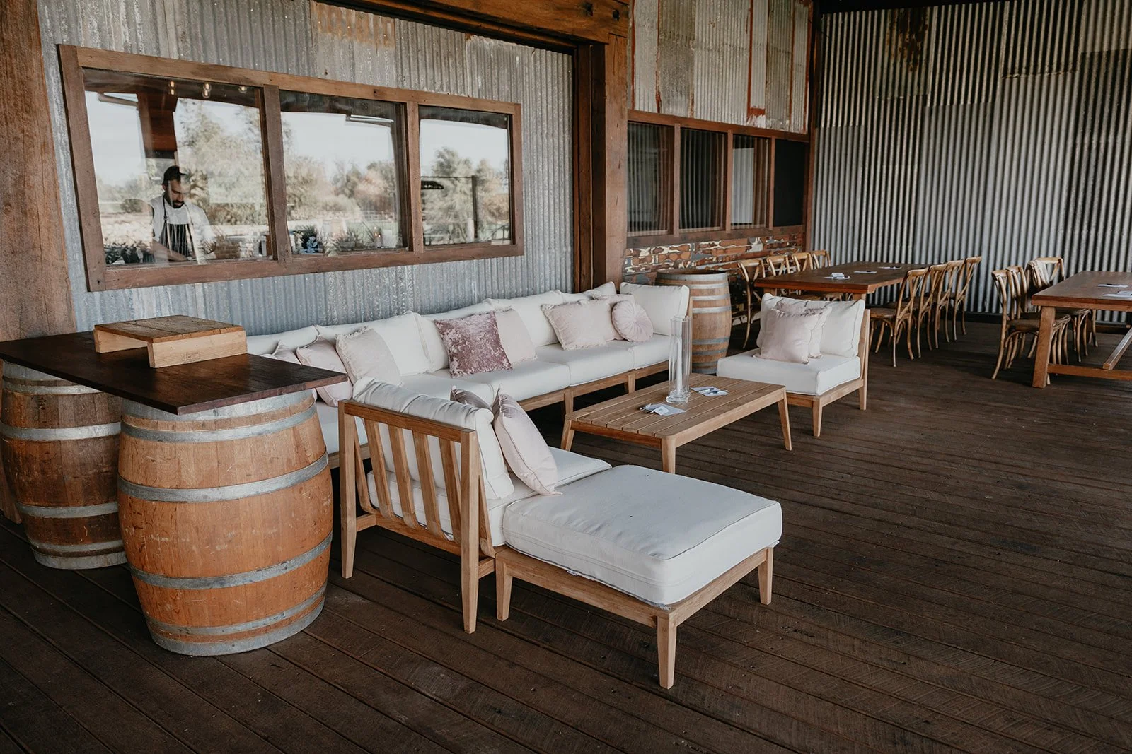 Veranda with wooden floorboards, a white sofa with pink and cream throw pillows, wooden tables, and chairs. Barrels used as side tables, a large window reflecting a chef in the kitchen, and corrugated metal walls.