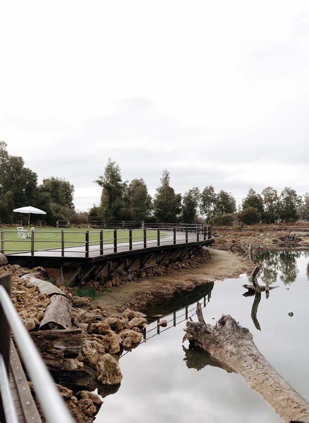 A wooden boardwalk alongside a body of water with rocks and fallen logs, leading to a grassy area with a white umbrella and chairs, surrounded by trees under an overcast sky.