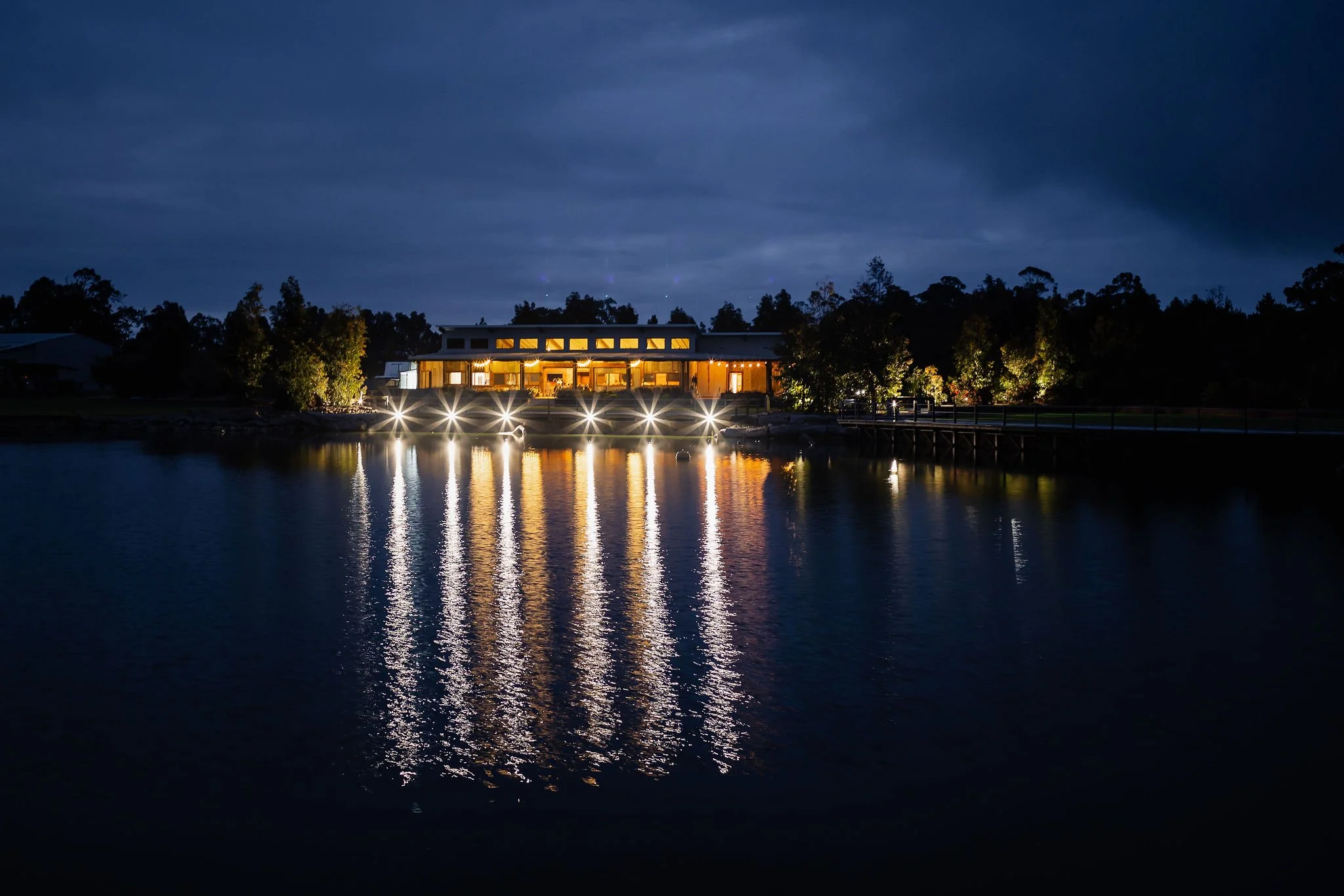 Nighttime view of a lit building by a body of water, with reflected lights on the water's surface, surrounded by trees and a cloudy sky.