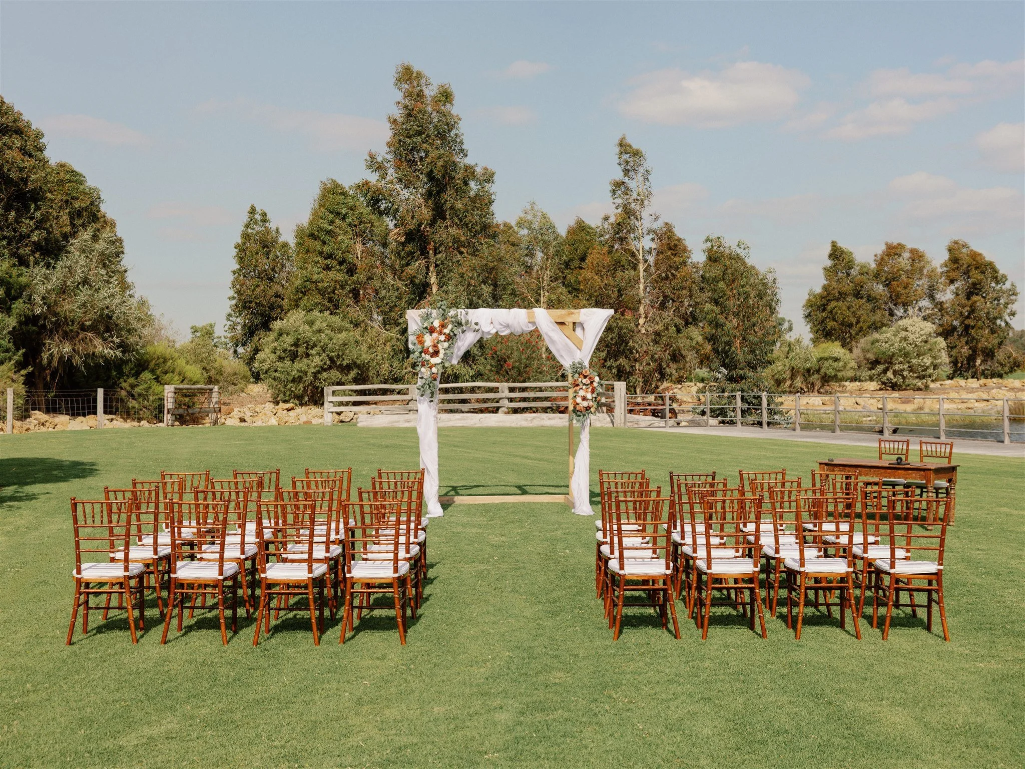 Outdoor wedding ceremony setup with chairs arranged in front of an arch decorated with flowers and white fabric, on a grassy field with trees and a blue sky in the background.