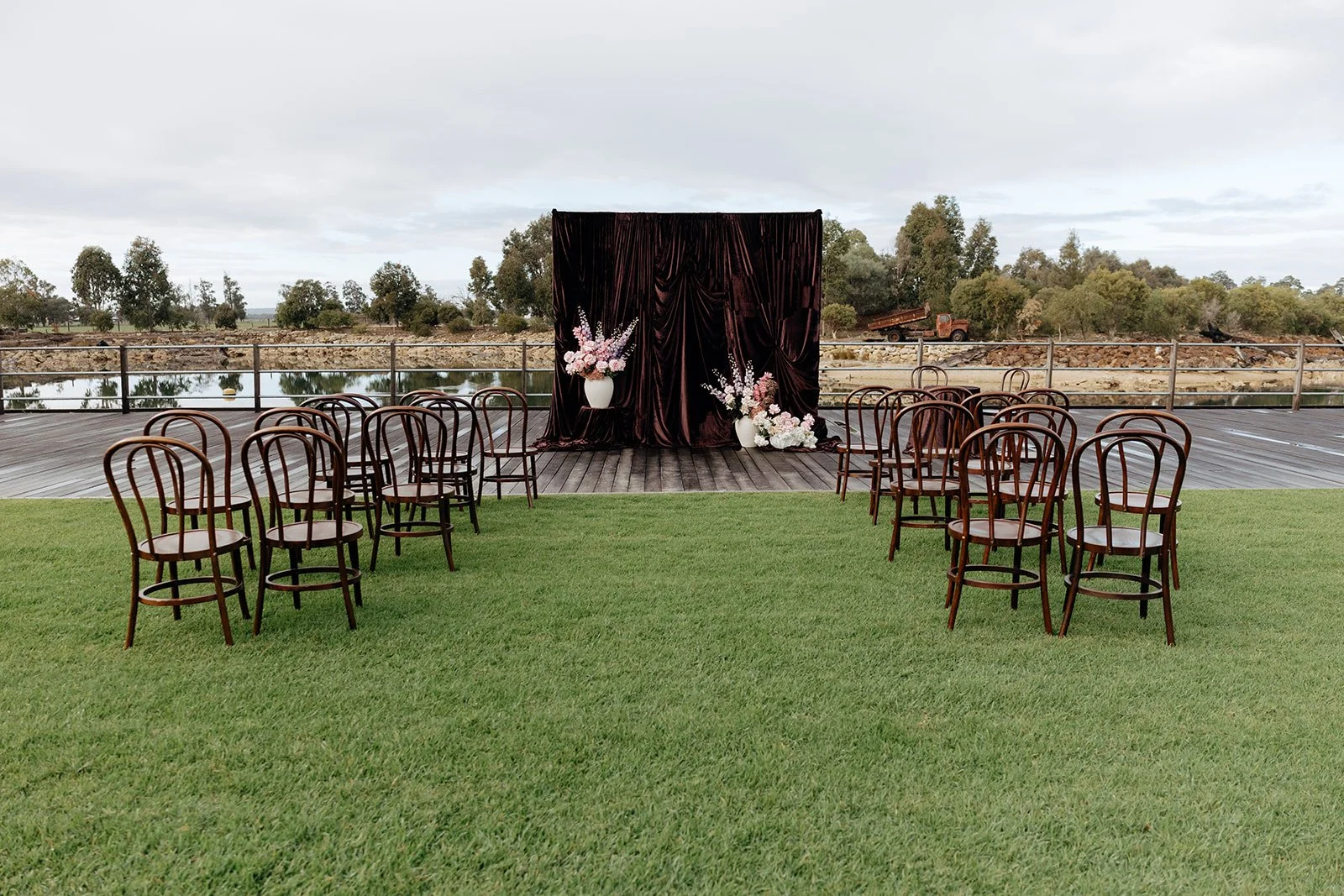 Outdoor wedding ceremony setup with chairs facing an altar draped with dark fabric and decorated with pink and white flowers near a body of water and trees.