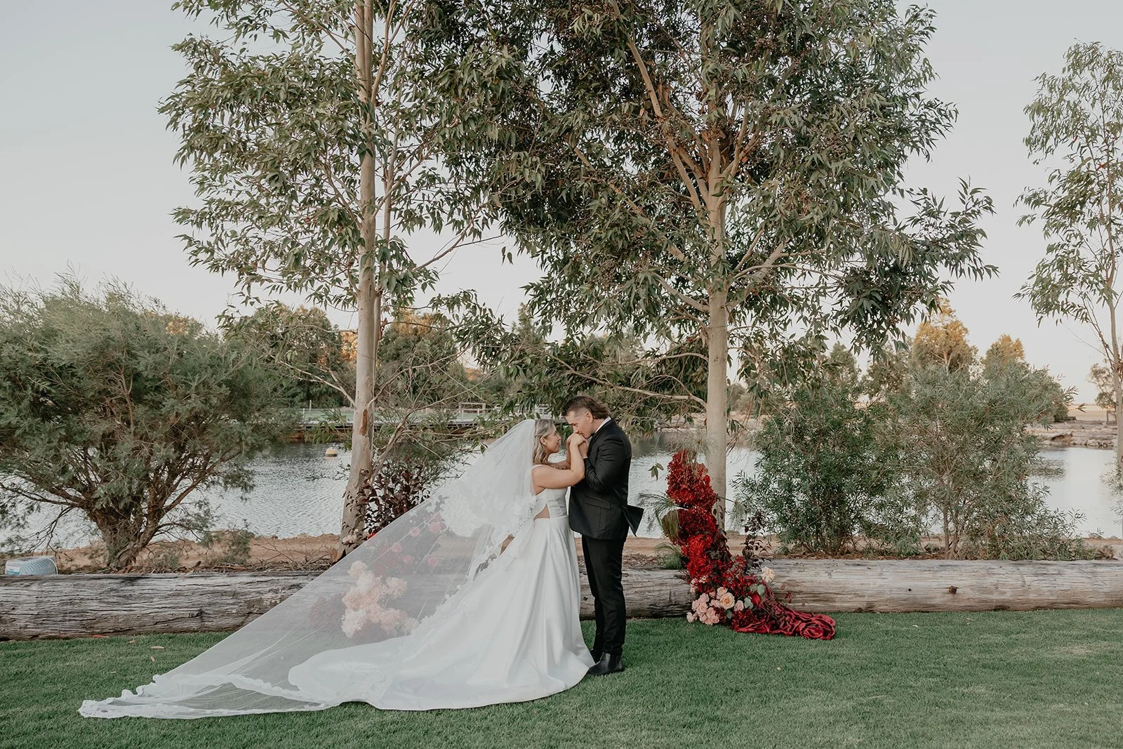 Bride and groom holding hands and touching foreheads during wedding ceremony outdoors near lake, with trees and floral arrangement in background.