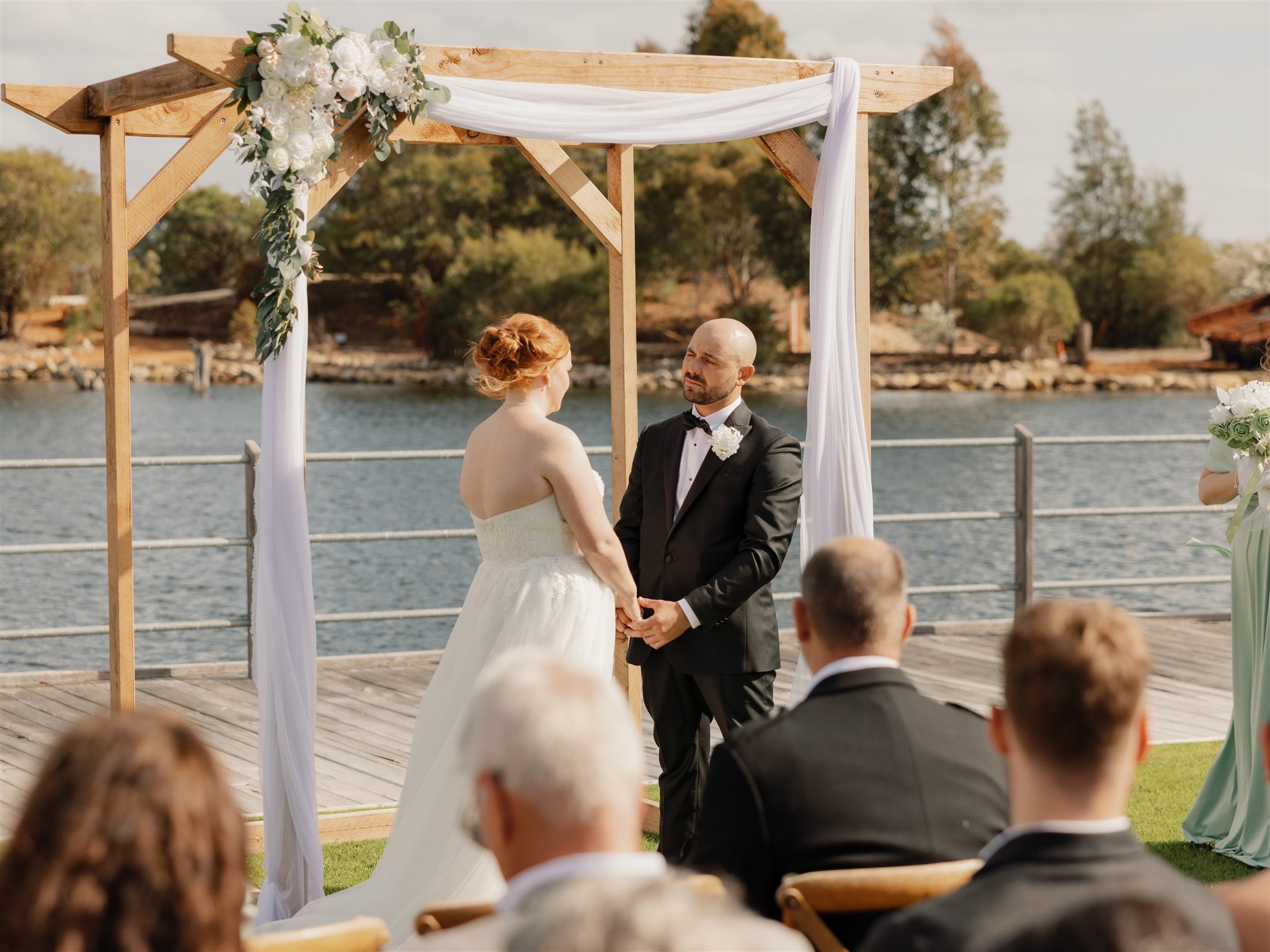 A couple gets married outdoors by a lake, standing under a wooden arch decorated with white flowers and fabric, with guests seated in front of them.