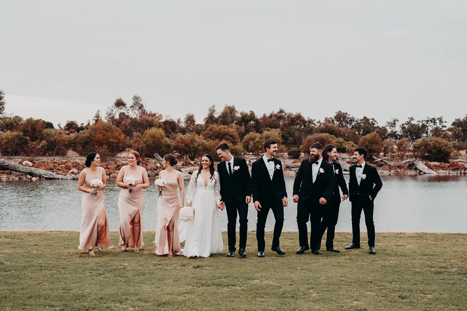 A group of wedding party members standing on grass near a body of water with trees and rocks in the background. The bride is wearing a white dress, and the groom and groomsmen are in black tuxedos. The bridesmaids are in blush-colored dresses, holding bouquets, and standing on the left side of the group.