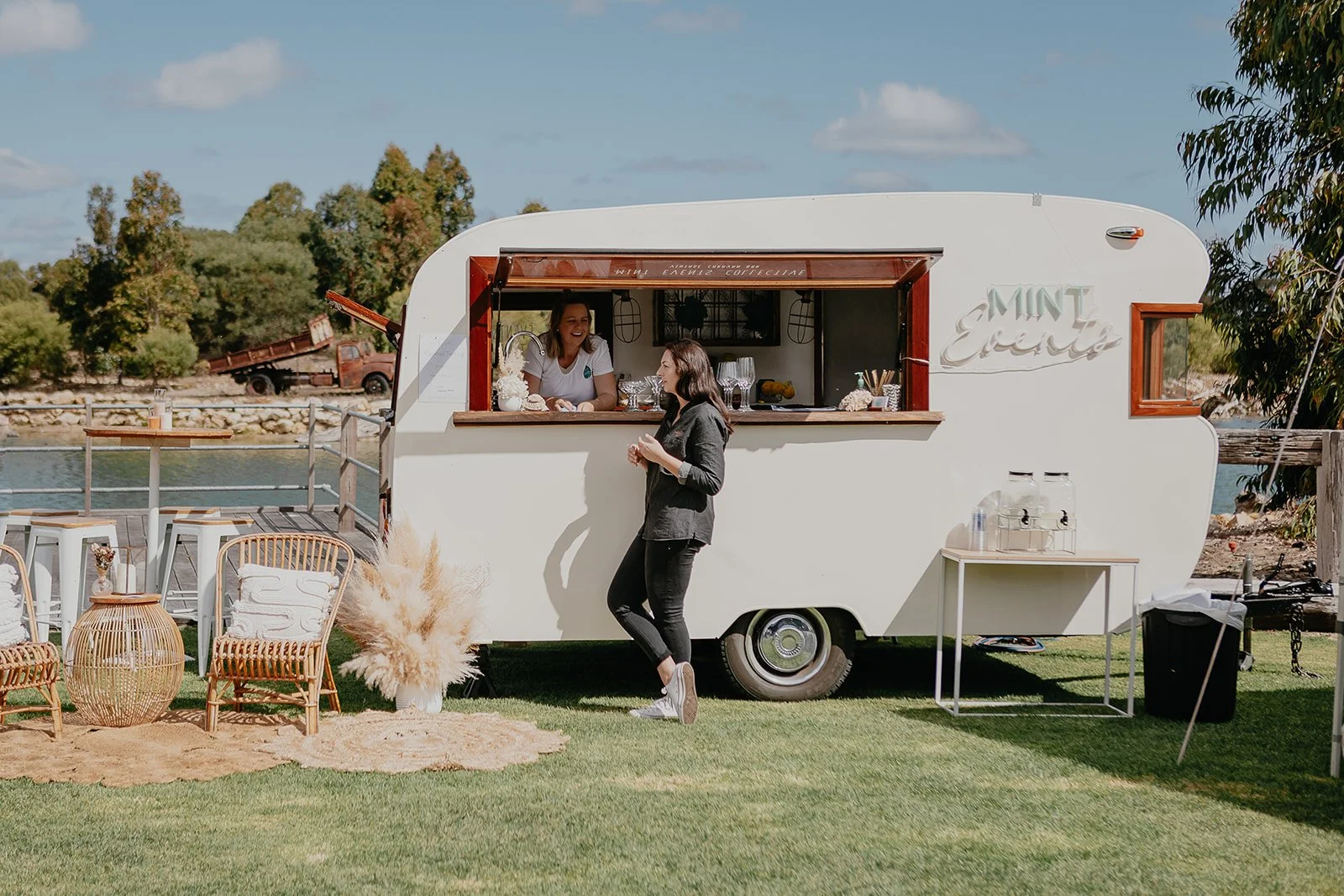 A white mobile food trailer labeled 'Mint Events' is parked outdoors near a river, with a woman standing outside talking to a smiling woman inside the trailer. The outdoor setting includes chairs, a small table with drinking water dispensers, and decorative plants.