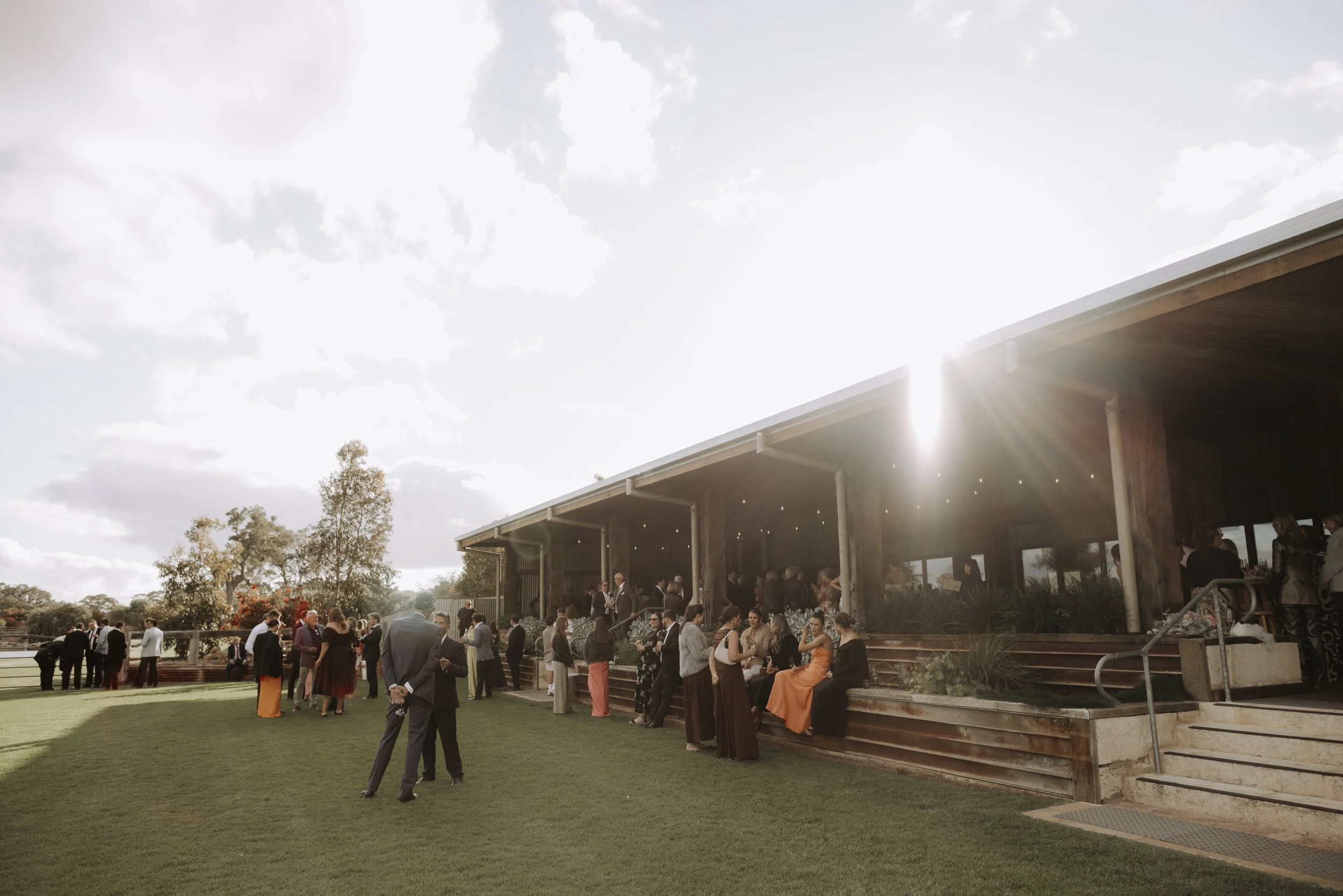 People gathered outdoors at a social event with a modern wooden pavilion, sitting and standing on a grassy lawn with trees in the background, under a partly cloudy sky with the sun shining brightly.