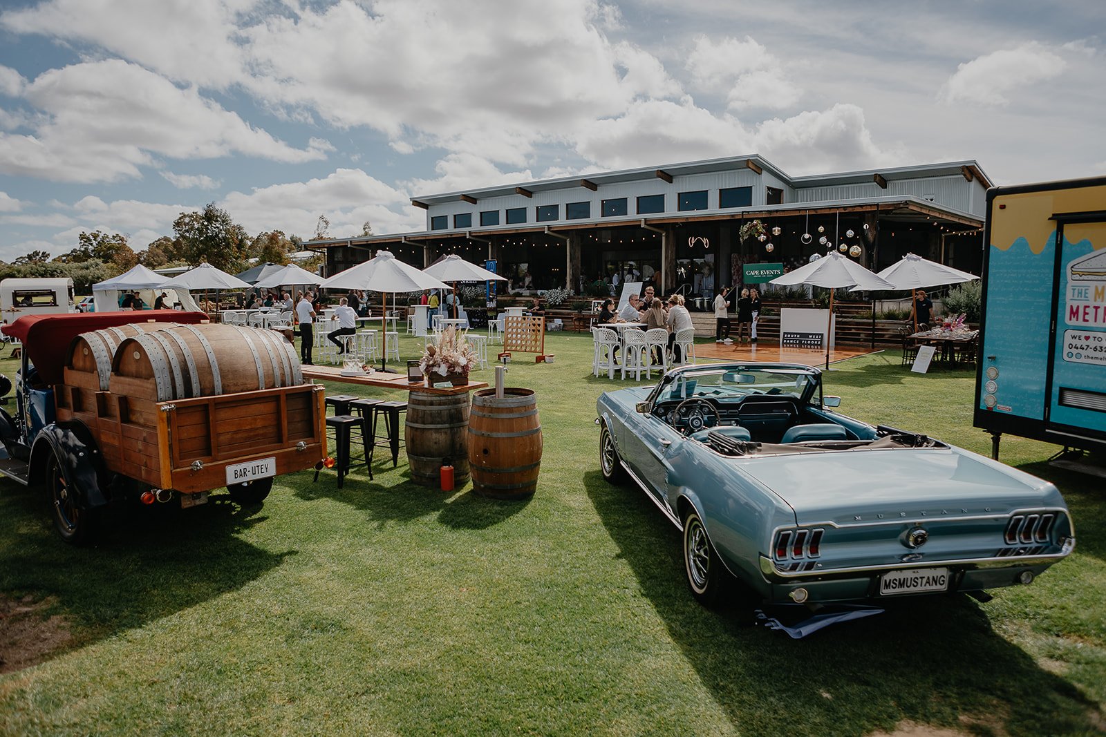 Outdoor event with vintage cars, barrel decor, tables, and umbrellas in front of a large building with cloudy sky in the background.