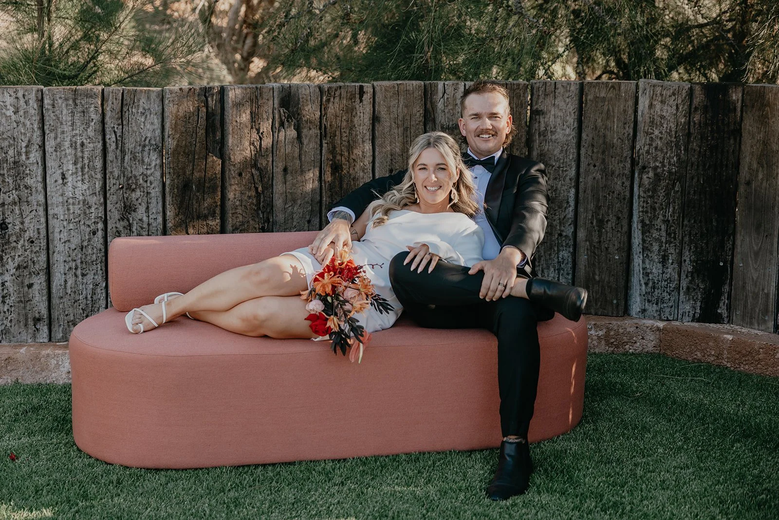 A couple sitting on a pink outdoor sofa with a wooden fence behind them. The woman is lying with her head on the man's lap, holding a bouquet of red, orange, and pink flowers. The man is sitting with one leg crossed over the other, wearing a black tuxedo with a bow tie, and has a big smile.
