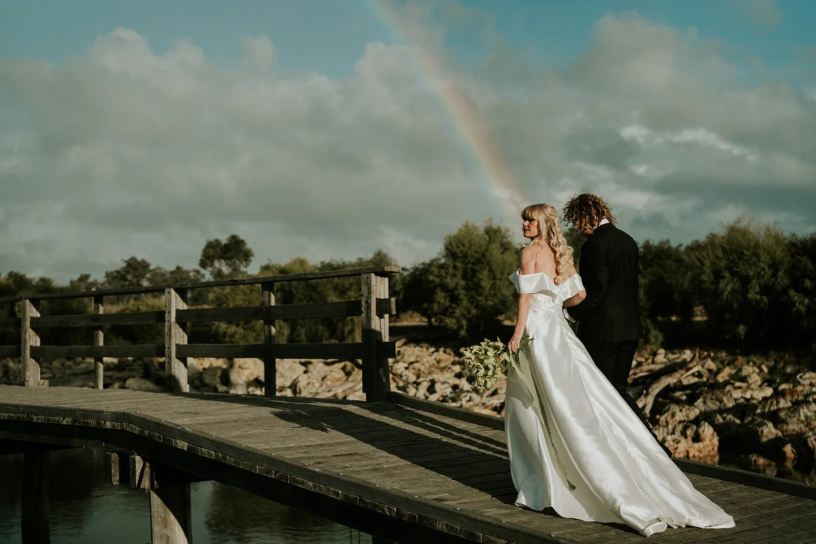 A bride and groom walking on a wooden dock by the water, with a rainbow and cloudy sky in the background, she holding a bouquet of flowers.