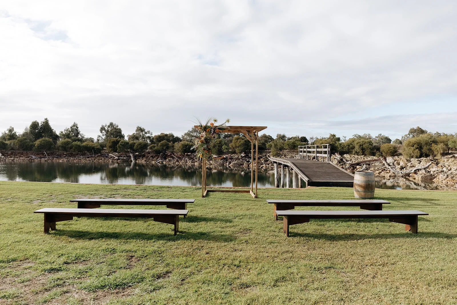 Outdoor wedding setup with benches facing a decorated arch by a body of water, with a wooden pier and barrel in the background, under a partly cloudy sky.