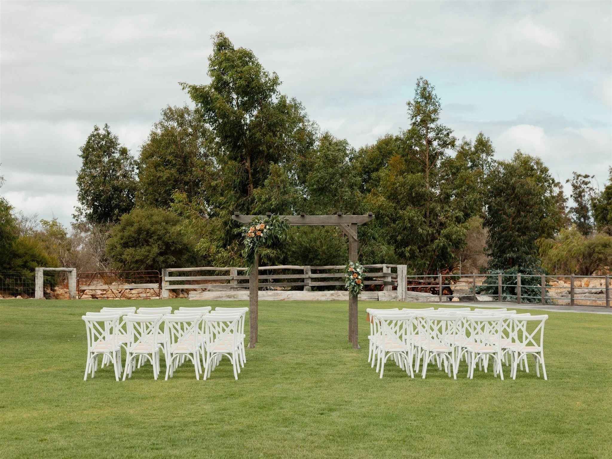 An outdoor wedding setup with a wooden arch decorated with flowers and greenery, surrounded by white chairs on a grassy lawn, with trees and a cloudy sky in the background.