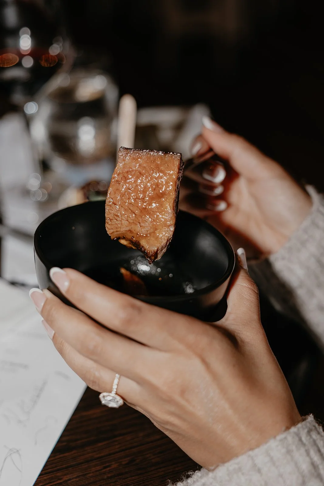 Person holding a piece of cooked meat with a fork over a black bowl, with a blurred background of a dining setting.