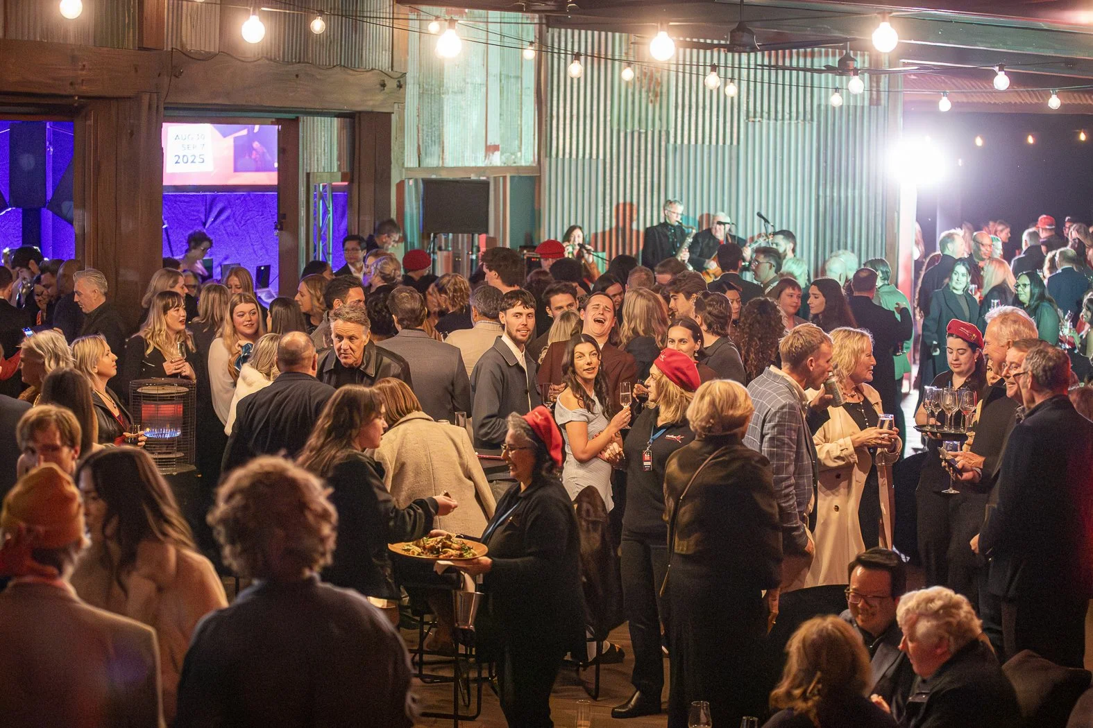 A lively indoor event with many people socializing, some holding drinks, in a rustic venue with string lights, a stage with musicians, and a large screen displaying the date September 7, 2025.