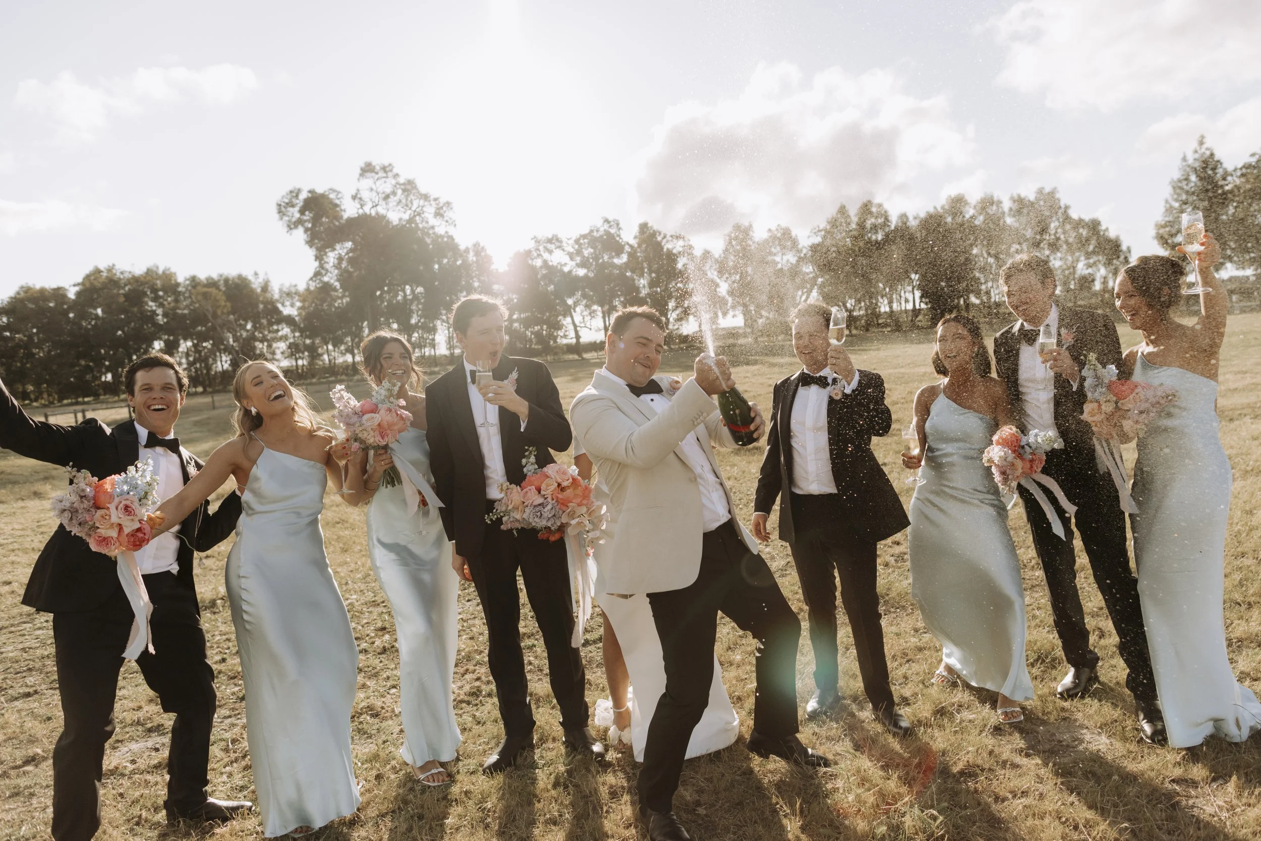 Wedding party celebrating outdoors in a field with champagne, smiling and holding bouquets, under a partly cloudy sky with the sun low on the horizon.